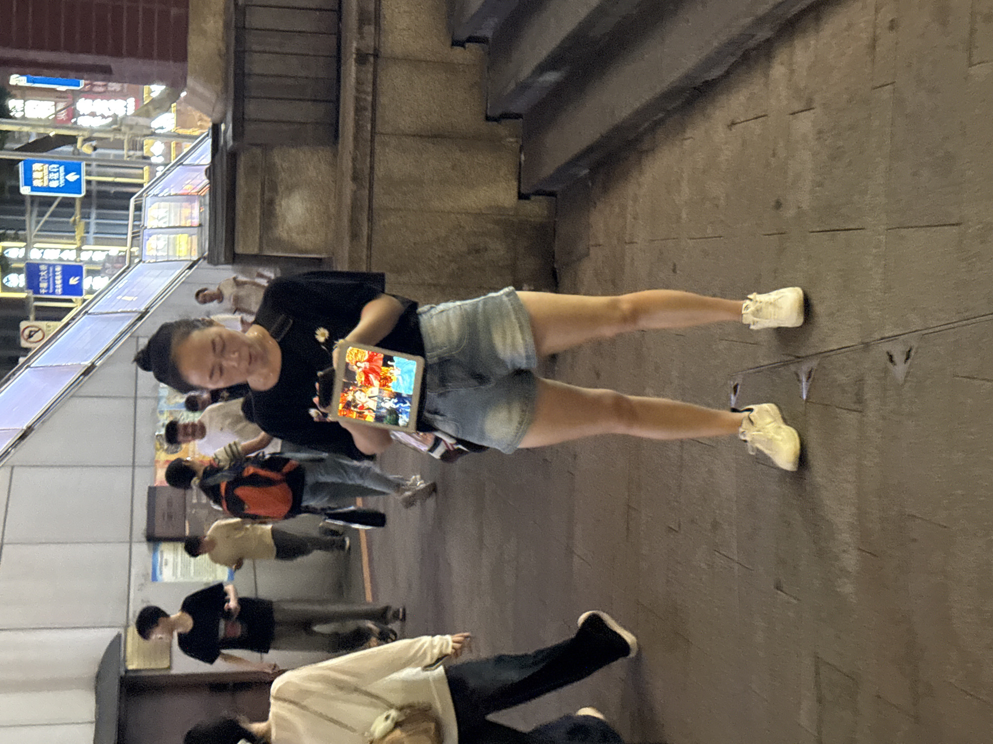 A woman in a black t-shirt and denim shorts stands on a paved walkway in Yuzhong District, China, holding a tablet displaying an image of colorful cartoon characters. She is wearing white sneakers and her hair is pulled up in a bun. In the background, several other people are walking, one wearing a red backpack, and an escalator with bright lighting is visible. The lighting suggests it is evening or nighttime. The environment appears to be an urban outdoor public space, possibly near a transit hub or commercial area.