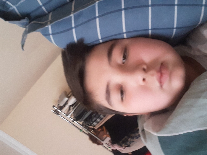 A young child lies on a blue plaid pillow, gazing languidly toward the viewer. Soft, rounded features and dark hair frame a serene face. To the left, a wall, and beyond, a clothing rack. The child's shirt, a light green and white striped garment, is partly visible. The shot is close-up, focusing primarily on the child's face.