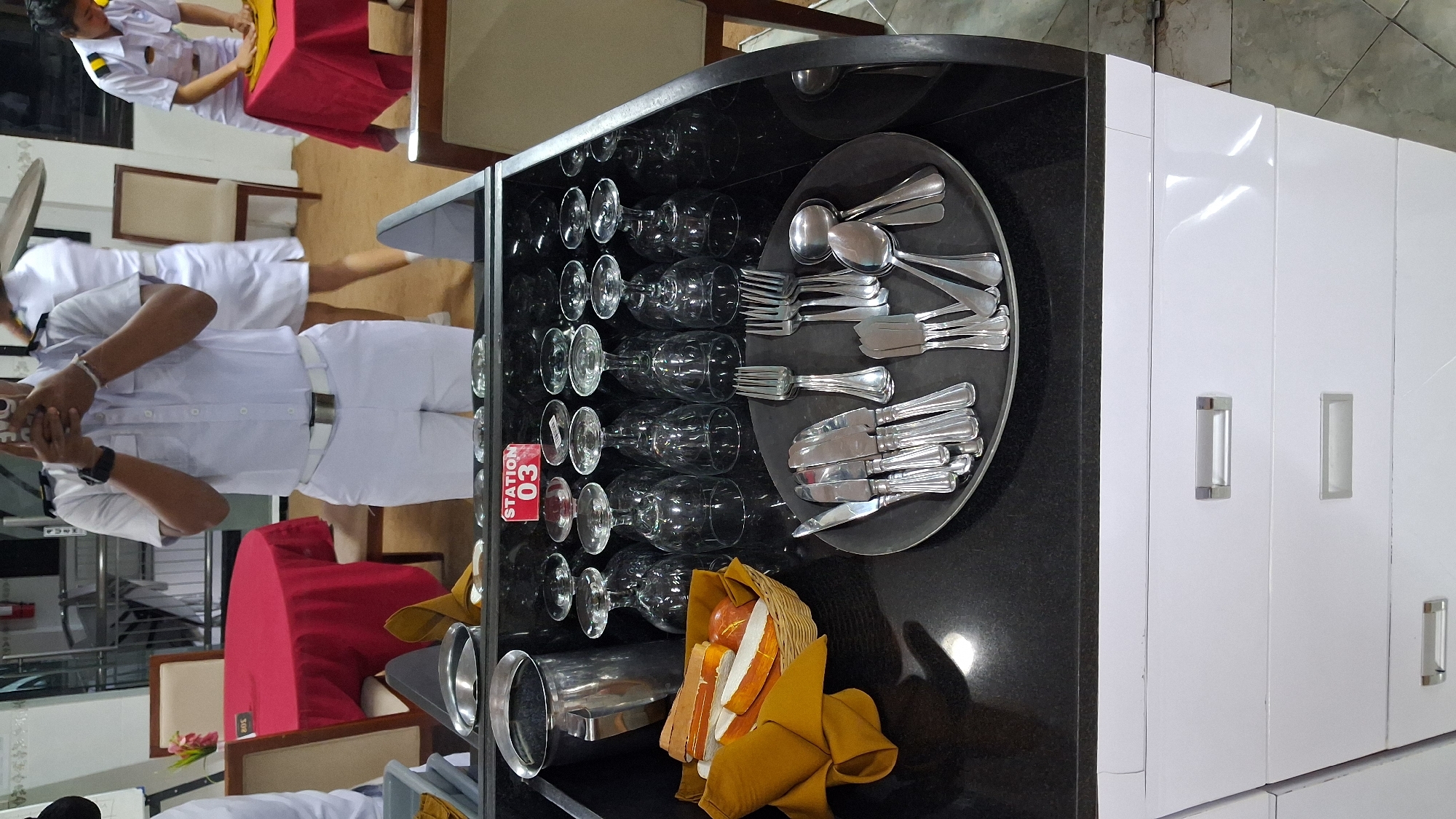 An overhead shot shows a black granite countertop, the top of a white cabinet with three drawers, and a portion of a person in the background wearing a white uniform. The countertop is organized with several rows of wine glasses, a woven basket filled with sliced bread, a large metal pitcher, and a dark gray circular tray containing a collection of silverware. A red sign with white text reading "STATION 03" is affixed to the front of the countertop. In the background, the person in the white uniform appears to be holding a phone. To the left and right of the countertop are glimpses of tables with red tablecloths and wooden chairs.