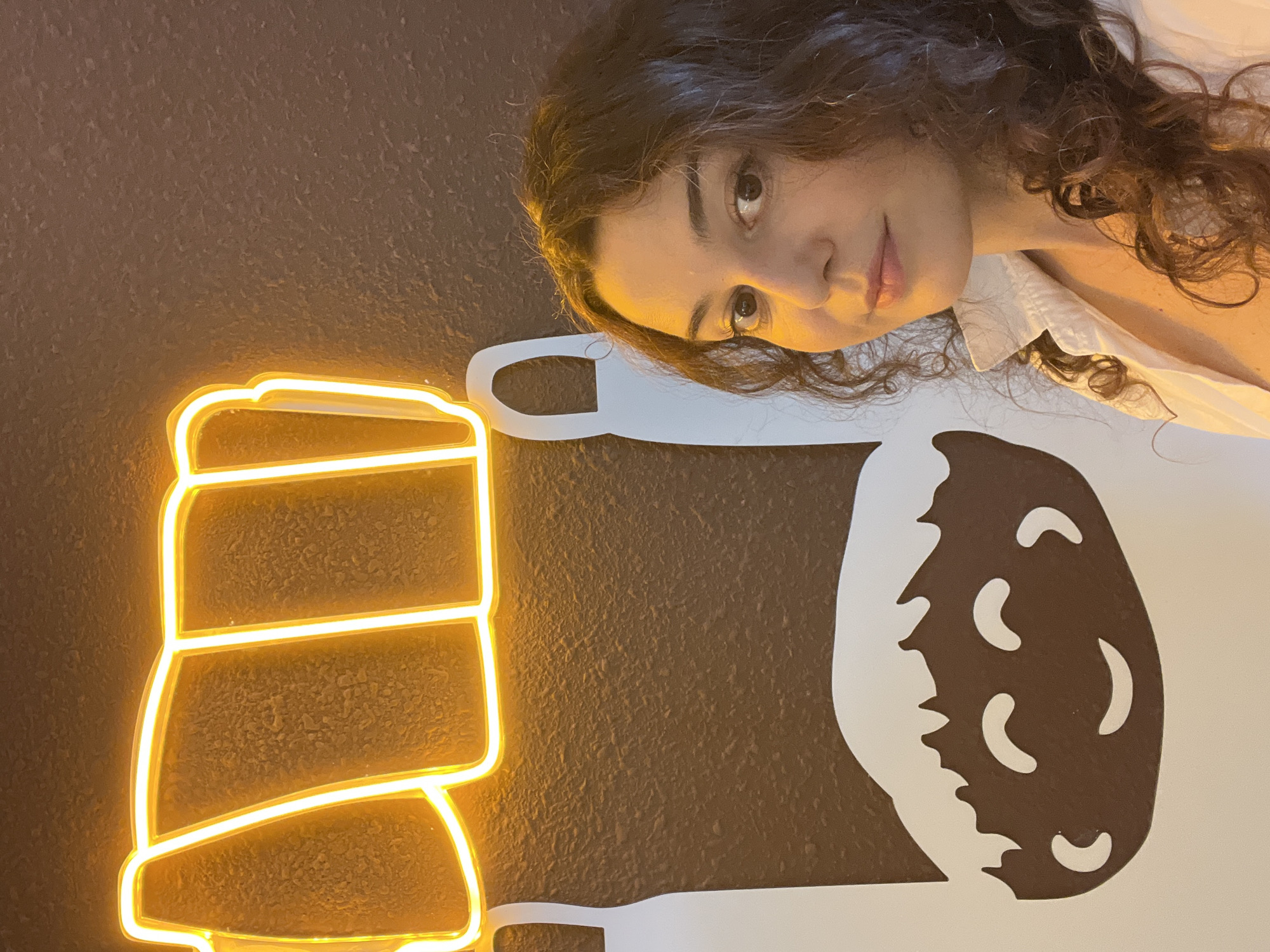 A young woman with curly brown hair and brown eyes, wearing a white collared shirt, is pictured from the chest up, posing for a selfie. She is standing in front of a dark brown textured wall. To her left, a neon sign emits a bright yellow-orange glow in the shape of what appears to be stylized bread loaves. Below the neon sign and partially behind the woman, there is a large graphic illustration on the wall. This illustration features two white shapes that resemble handles of a bag, with a brown cartoon face with a smiling expression and white hair details below them. The overall lighting in the image is warm, primarily from the neon sign, casting a yellow hue on the wall and the woman's skin.