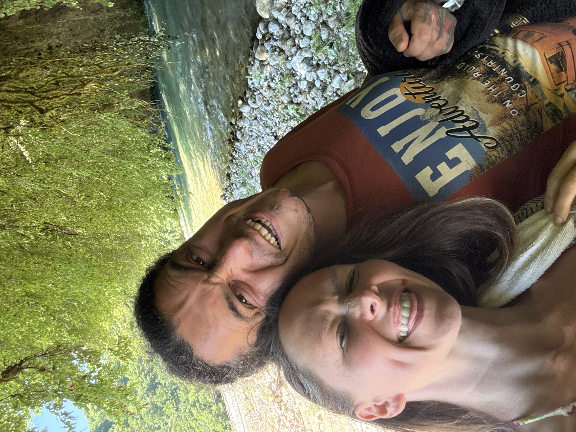 This is a close-up, low-angle, outdoor selfie of a man and a woman. They are positioned close together, with the man's face on the left and the woman's on the right. Both are looking towards the camera with exaggerated expressions, scrunching up their faces and showing their teeth as if laughing or making a funny face.
The man has dark, curly hair and a beard, and is wearing a rust-colored t-shirt with a graphic design. The woman has long, dark brown hair and is wearing a patterned top.
The background shows a river on the left and a rocky shore with greenery on the right, suggesting they are by a body of water in a natural setting. The sunlight appears bright, indicating it is daytime.
On the man's t-shirt, there is visible text in a yellow, stylized font. The words "ENJOY" are prominent, and below that, "ADVENTURE" and "ON THE ROAD" are partially visible, along with "COUNTRY."
The overall mood is playful and intimate, capturing a moment of shared amusement between the couple in an outdoor environment. The location context suggests they are in Abgara, Abkhazia.