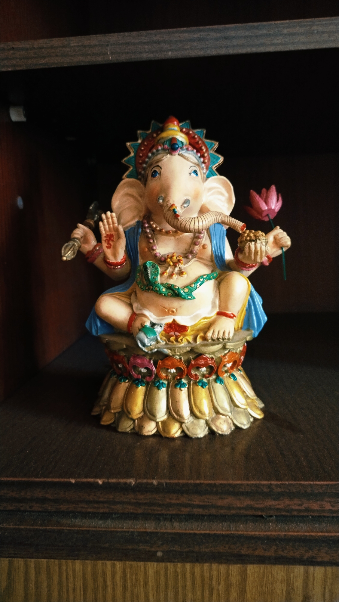 A Ganesha figurine is displayed on a dark wooden shelf. The figurine is intricately detailed and colorful, depicting the elephant-headed deity seated in a meditative pose. Ganesha is adorned with a blue sash, necklaces, and a green snake wrapped around his torso. He holds a lotus flower in his right hand and a ceremonial staff in his left. The base of the figurine is elaborately decorated with lotus petals in various shades of gold, yellow, and red, with blue and green accents. The background is a dark, shadowed area, likely a shelf or cabinet. The image was taken in Horishni Plavni, Ukraine.