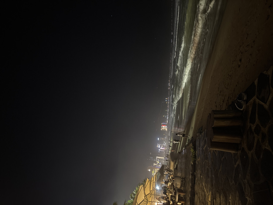 The image depicts a nighttime scene at a beachfront in Ngũ Hành Sơn Ward, Vietnam. The foreground features a dark-colored irregular stone-paved walkway or patio area. On this pavement, a cluster of three cylindrical wooden stools or bollards is visible, alongside a pair of dark-colored flip-flops. To the left, an outdoor dining or lounge area is established, characterized by multiple light-colored, open patio umbrellas illuminating tables and chairs where several indistinct figures are seated, appearing to socialize or dine. Behind this area, some dark vegetation, possibly palm trees, is discernible.
The mid-ground transitions to a sandy beach extending horizontally, where three to four individuals, likely children or young adults, are observed walking or standing near the waterline. To the right, the dark ocean with illuminated breaking waves rolls onto the shore. The background displays a long stretch of distant city lights along the coastline, indicating a densely built urban area with visible high-rise buildings. The sky above is dark and appears slightly hazy, with no clear celestial bodies visible. Artificial lighting from the establishments and distant city sources casts illumination across parts of the beach and waves.