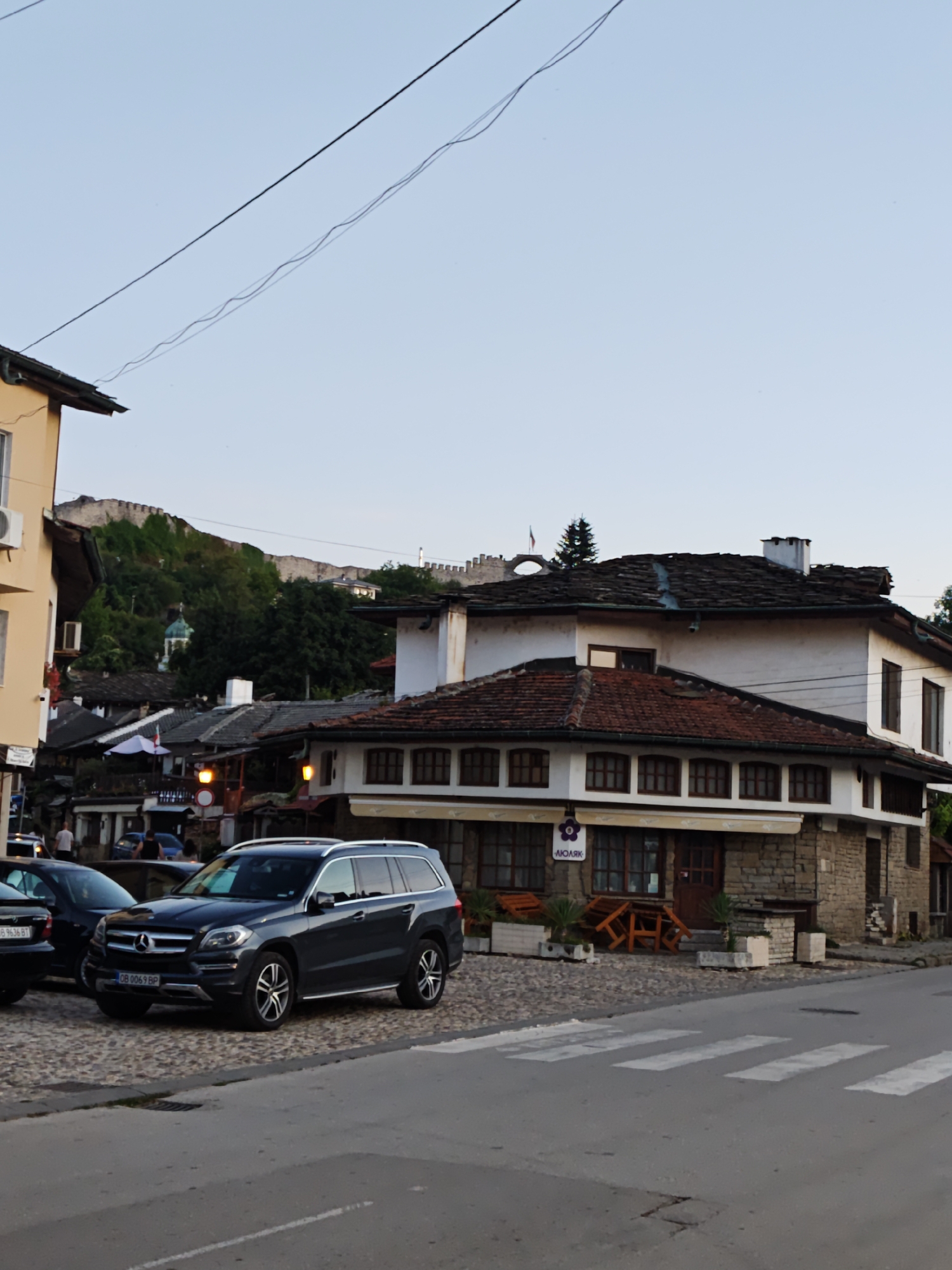 The image depicts a street scene in the city of Lovech, Bulgaria. In the foreground, a dark gray Mercedes-Benz GL-Class SUV is parked on a cobblestone street. To the left of the SUV, the rear of a dark-colored sedan is visible. A pedestrian crossing is marked with white stripes on the asphalt road in the lower right portion of the image.
Behind the parked vehicles, a two-story building with a stone base and white-painted upper walls dominates the mid-ground. This building features a red-tiled roof with visible shingles and multiple dark-framed windows. A sign with Cyrillic lettering and a floral logo is affixed above the ground-floor windows. Outdoor seating areas with wooden tables and chairs are visible in front of this building.
Further in the background, a cluster of older buildings with dark tiled roofs line the street. A Bulgarian flag is displayed on a pole above one of these structures. In the far background, the imposing walls of Lovech Fortress are perched atop a hill, partially obscured by lush green trees and vegetation. A small, green-domed structure, likely a church or chapel, is nestled amongst the trees below the fortress. The sky is clear and pale blue, suggesting it is either early morning or late afternoon. The overall impression is a historic European town street, with a blend of traditional architecture and modern vehicles.