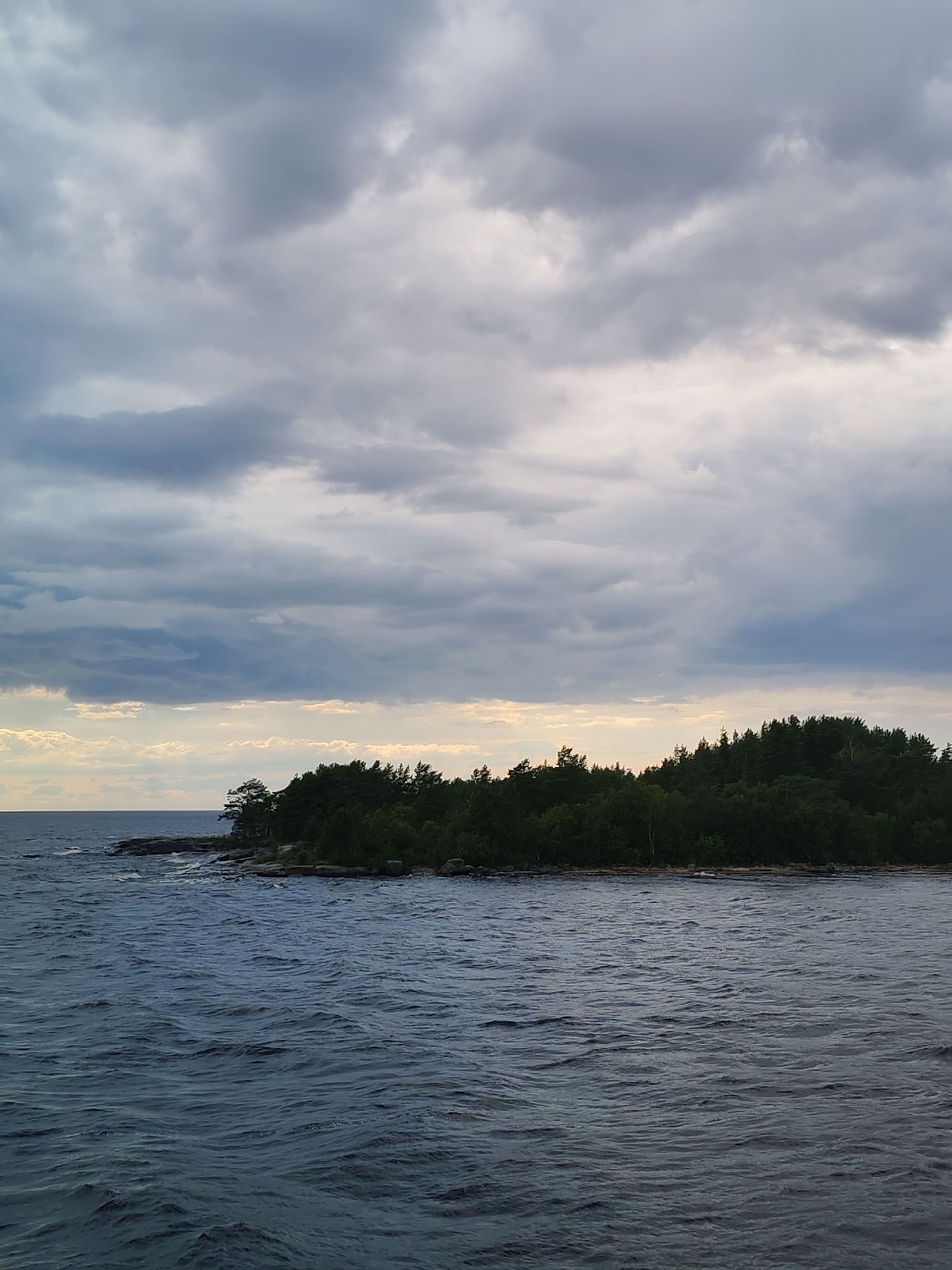 A wide shot captures a dramatic, overcast sky dominating the upper portion of the frame, with layers of gray and white clouds creating a textured ceiling. Beneath the clouds, a sliver of pale yellow light pierces the horizon, suggesting either sunrise or sunset. The middle ground features a densely wooded island, its dark green trees forming a solid mass against the sky. Rocky outcroppings are visible along the island's shoreline, jutting into the dark blue water. The foreground is filled with the choppy surface of the water, showing small waves and ripples reflecting the muted light. The scene evokes a sense of natural beauty and tranquility. This image appears to be of the shoreline near Shalsky, Russia.