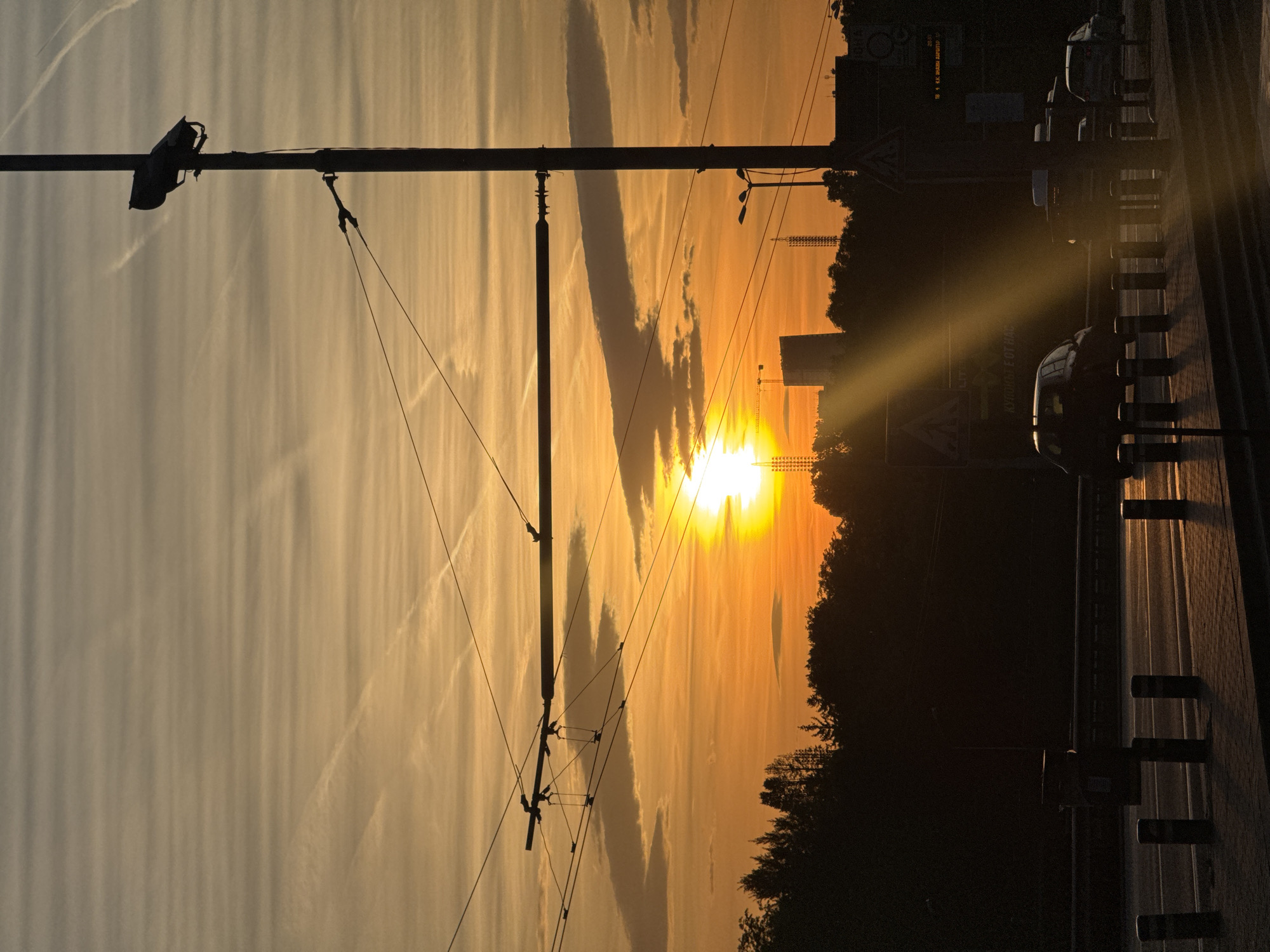 The image captures a sunset in Sofia, Bulgaria. The sun is positioned low on the horizon, obscured by trees and buildings. Silhouetted against the bright sky are electrical poles and wires. A road with parked cars runs along the right side of the frame, with a curb and barrier visible. The sky is a mix of orange and yellow hues, with some clouds present. Vapor trails from aircraft are visible. The time of day is dusk. There are trees to the right of the image.
