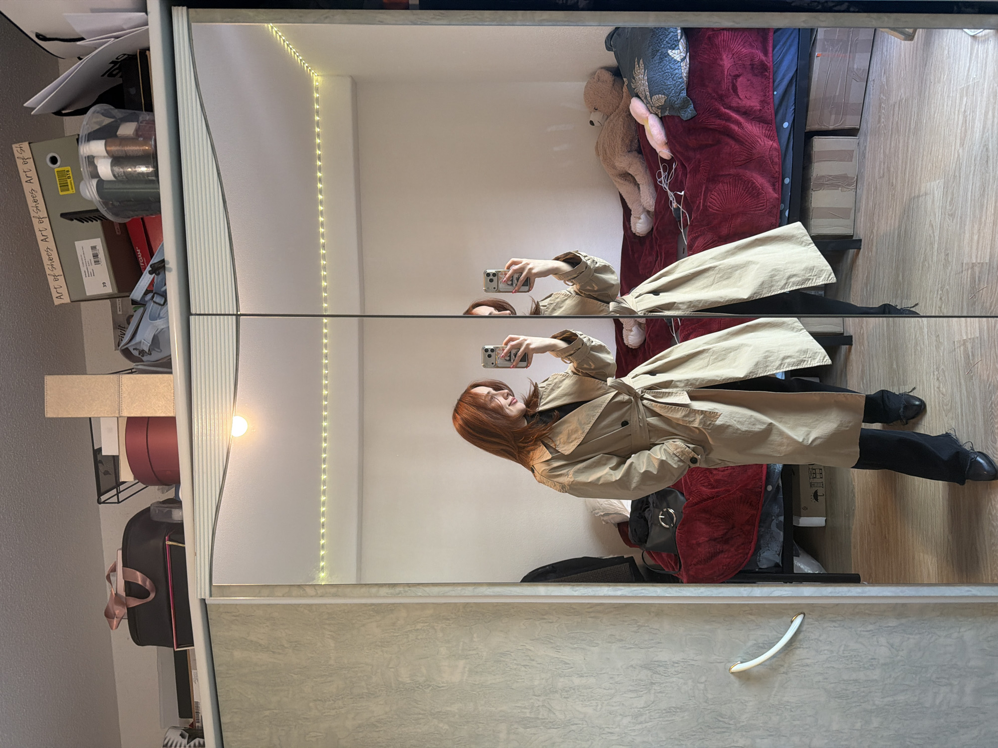 An indoor scene in a residential dwelling located in Strasbourg, France, is depicted. The image is a full-body mirror selfie taken by a single female subject.
The subject, with reddish-brown hair styled loosely, is reflected standing upright, facing slightly right, while holding a silver-colored smartphone in both hands to capture her reflection. She wears a light tan, double-breasted trench coat with a tied belt at the waist, a black turtleneck shirt, black trousers with frayed hems, and black pointed-toe flat shoes. Red nail polish is visible on her fingers.
The reflection reveals a bedroom setting. Behind the subject, a bed with a dark red quilted duvet cover is visible. On the bed, a large light brown teddy bear lies on its side, alongside a smaller pink plush toy and a dark blue cushion with a light-colored pattern. Tangles of white cords are present on the duvet. A black handbag with a prominent silver circular detail rests on the bed. The wall behind the bed is white, and a string of warm white LED fairy lights runs horizontally across the top portion of this wall. A segment of a black metal bed frame is visible, and two taped cardboard boxes are stored beneath the bed. The flooring is light wood laminate with visible grain.
Above the mirrored cabinet, which also serves as the reflective surface, various items are stored: a black duffel bag with a pink handle, a red cylindrical box, a black wire storage basket, a light blue bag, a clear plastic container with small bottles, and several cardboard boxes, one explicitly labeled "Art of Shoes" with the number "39" and a "BTB" sticker. The cabinet itself features a non-mirrored left panel with a textured light gray/beige surface and a white curved handle with gold tips. A single light source, emitting a warm yellow glow, is affixed above the mirrored panels.