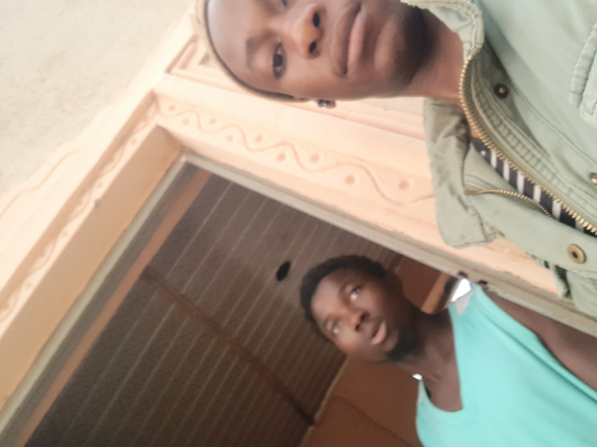 Two African men are posing for a selfie, possibly at the city of Dutsinma A, Nigeria. The man closer to the camera is wearing a green jacket with a black and white striped shirt underneath. The second man, positioned behind and below, is wearing a light blue tank top. They are standing in a doorway, the doorframe is light orange and has a decorative pattern. The interior behind them seems to be a metal-roofed structure.
