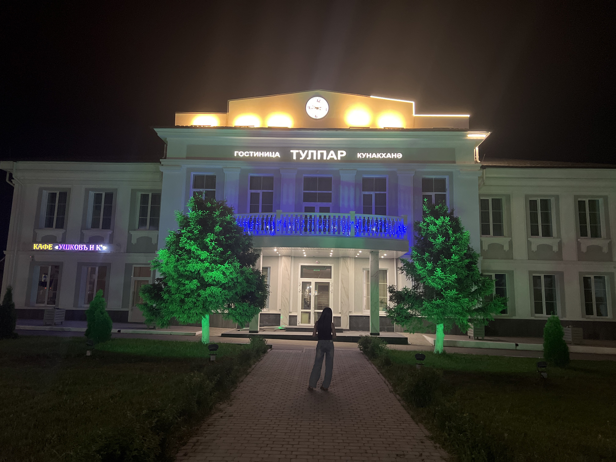 At night in Mendeleyevsk, Russia, an unidentified person with long dark hair stands on a paved path, facing away from the camera and toward a building. The building is identified as "гостиница ТУЛПАР КУНАКХАНӘ" (Hotel Tulpar). The building also has a sign for a cafe, "КАФЕ «УШКОВЪ И К»" (Cafe Ushkov & K). The front of the building is illuminated with blue lights strung along a balcony. Two trees flank the path leading to the building, illuminated in green. A clock is centered on the top of the building. The sky is dark, indicating it is nighttime.