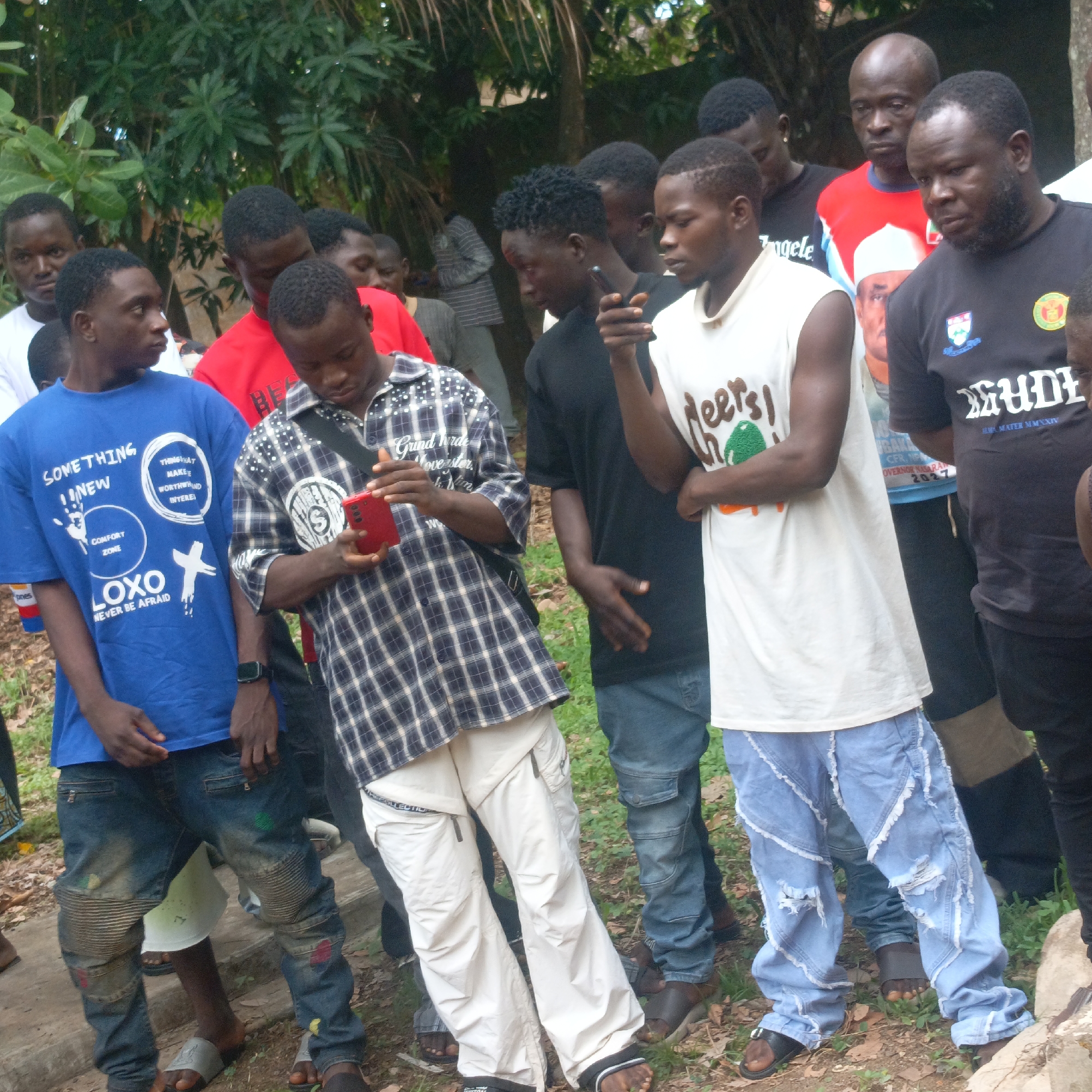 The image shows a group of approximately 14 African men standing outdoors in Andaha, Nigeria. Most are dressed casually in t-shirts and jeans or casual trousers, with several wearing sandals. Some are looking at their mobile phones. One man is wearing a t-shirt promoting a political candidate, C.F.R. NDIGBARA, Governor of Nasarawa in 2027. The environment is somewhat overgrown with grass and trees in the background.