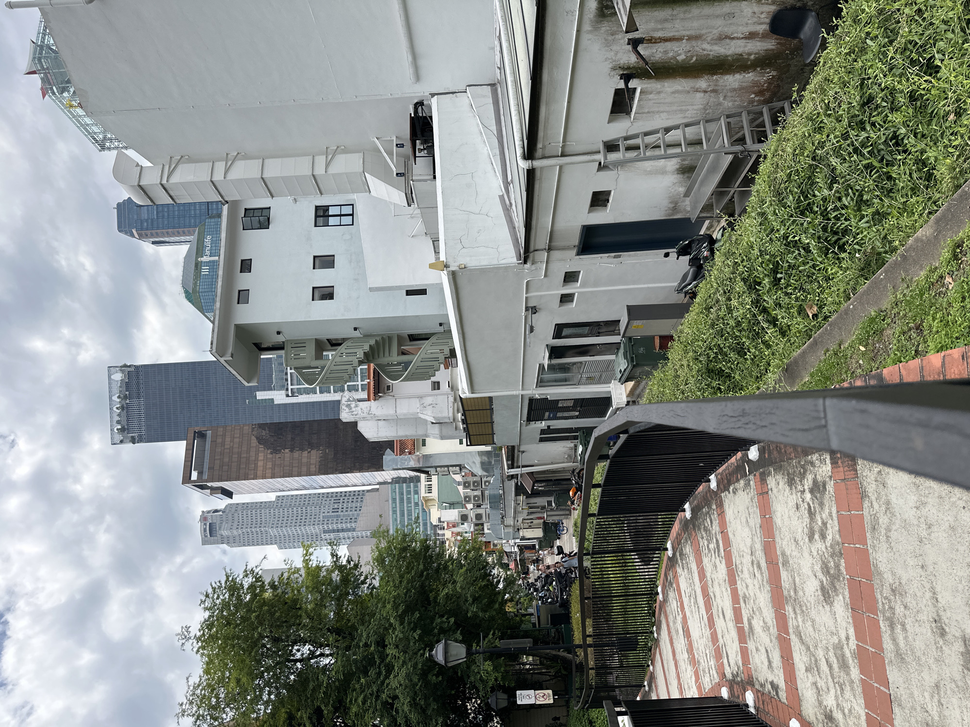 The image depicts a street scene in Singapore, possibly in the Chinatown or Kampong Glam area, known for its blend of old and new architecture. In the foreground, a winding brick pathway with a black metal railing leads downwards. To the right, a white building with multiple windows and an external spiral staircase is visible. Beyond this, more white buildings with various air conditioning units are present. In the background, several modern skyscrapers pierce the cloudy sky, contrasting with the lower-rise, older structures. A few motorbikes are parked along the street, and a lone black chair sits amongst dense green foliage to the right. The overall atmosphere is one of an urban environment with historical and contemporary elements coexisting.