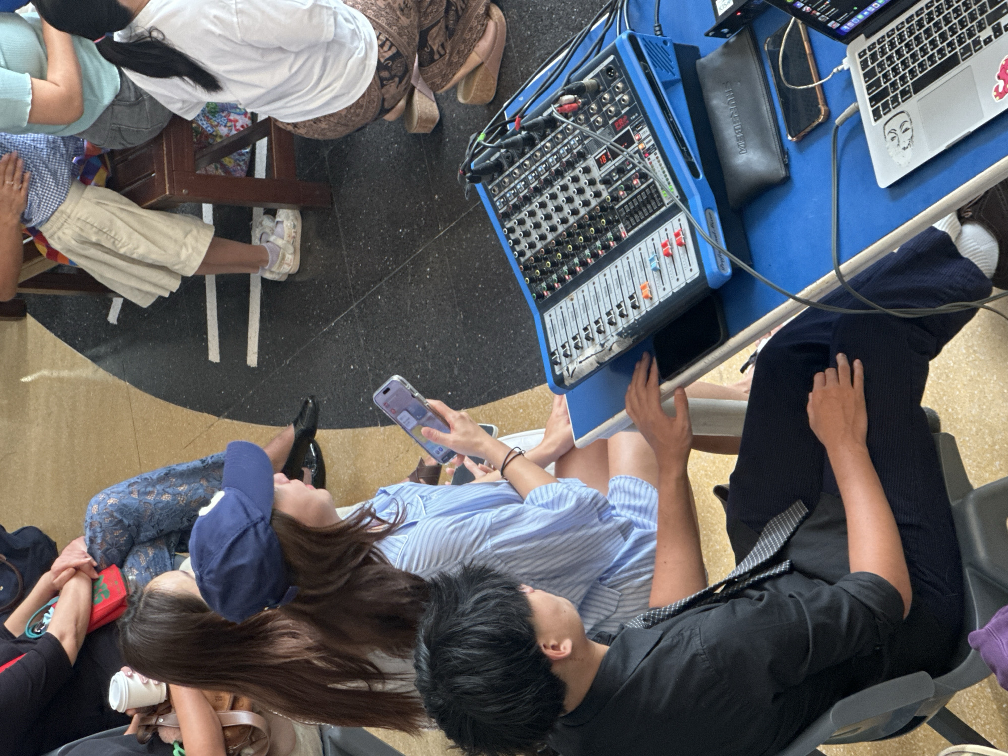 This image shows a group of people gathered around a blue table with a sound mixer and a laptop. A young man in a black shirt and tie is seated to the left, looking towards the mixer. A young woman in a blue and white striped shirt and a blue baseball cap is seated in the center, holding a smartphone. Behind them, other individuals are partially visible, some seated and others in the background. The environment appears to be indoors, with a dark floor and light-colored walls. The scene is likely at a public venue or event in Kathu, Thailand.