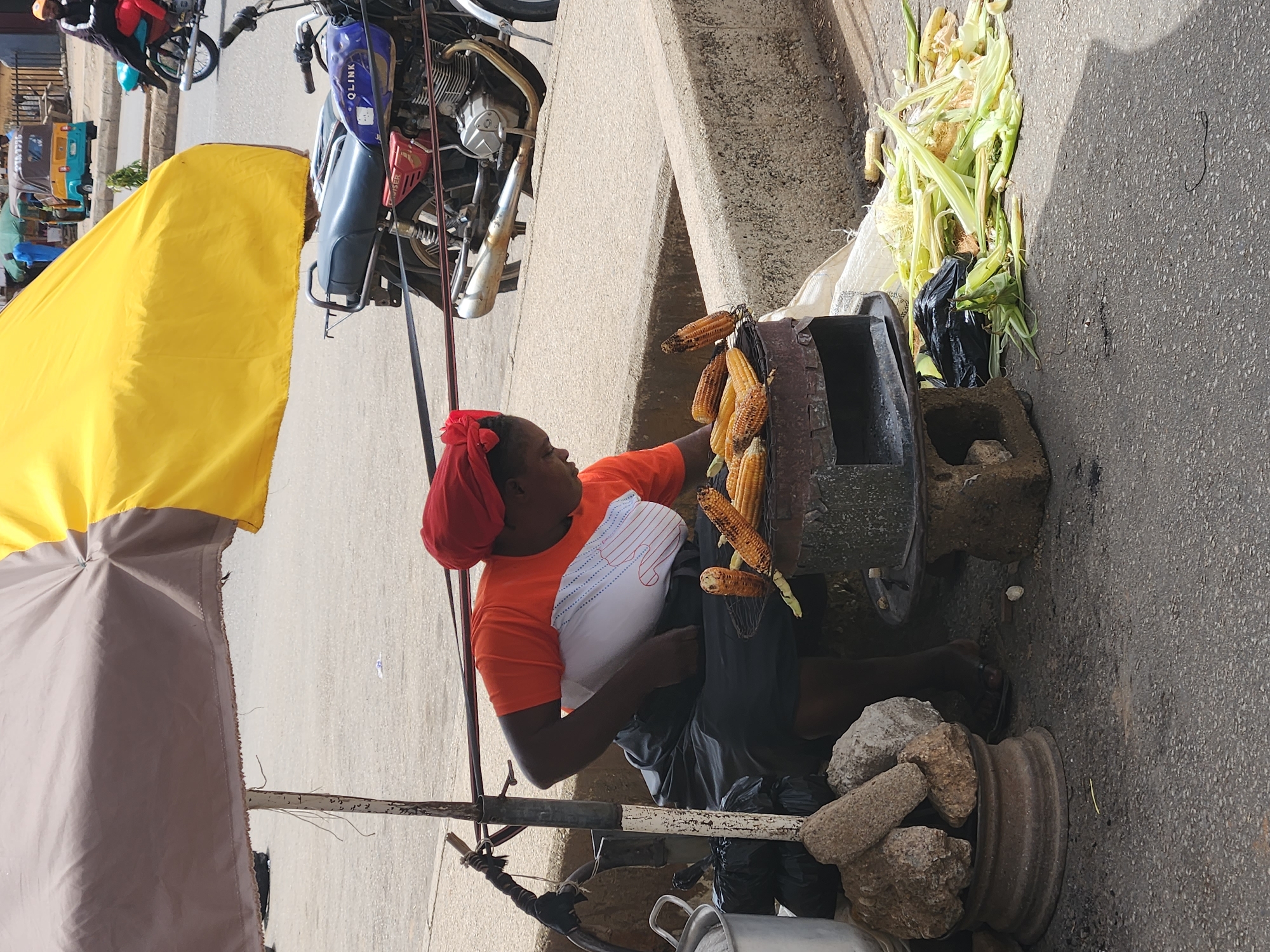 The image shows a woman selling grilled corn on a street in Kabong, Nigeria. She is sitting next to a grill with several ears of corn roasting on it. She has a red headscarf and is wearing an orange shirt. A large pile of fresh corn, still in their husks, sits to the right of the grill. She is positioned underneath a makeshift awning made of yellow and grey fabric. A motorcycle is parked nearby. The street is asphalt and there is a concrete curb. Stones are piled in a rusted wheel rim.
