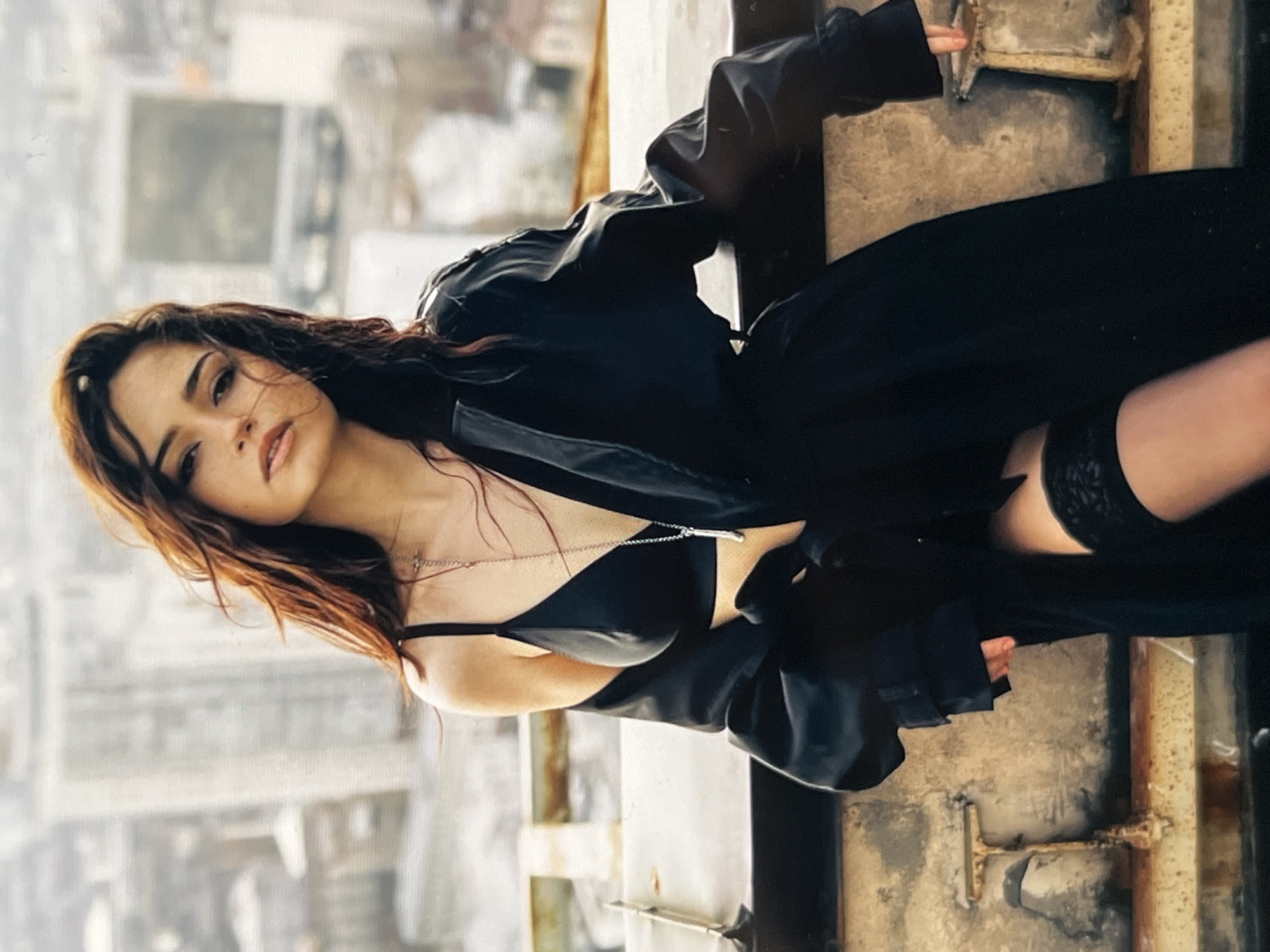 A young woman with long, dark brown hair is positioned on what appears to be a rooftop in an urban setting. She is wearing a black bra, a black trench coat, and black thigh-high stockings. A silver necklace with a pendant hangs around her neck. She is leaning against a metal barrier, with a blurred city skyline visible in the background. The lighting suggests an overcast day.