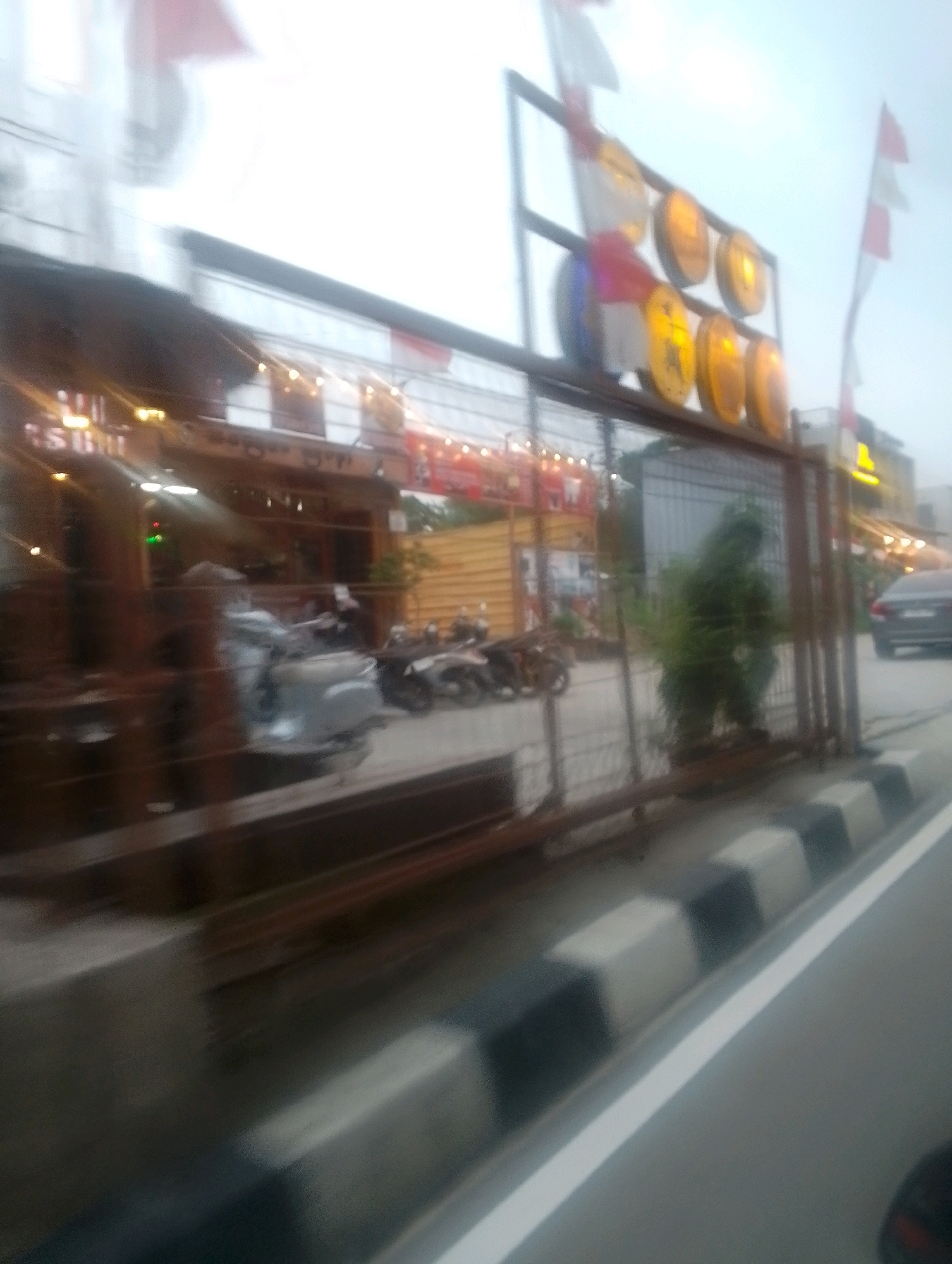 The image shows a blurred street scene in Depok, Indonesia. In the foreground, a black and white striped curb runs along a road. To the left, a building with lights and signage is visible, possibly a restaurant or shop. Several motorcycles are parked in front of the building. A fence separates the sidewalk from the building area. In the background, more buildings and street lights can be seen. A dark-colored car is parked on the right side of the road. The overall impression is that of a busy urban environment, likely in the evening.