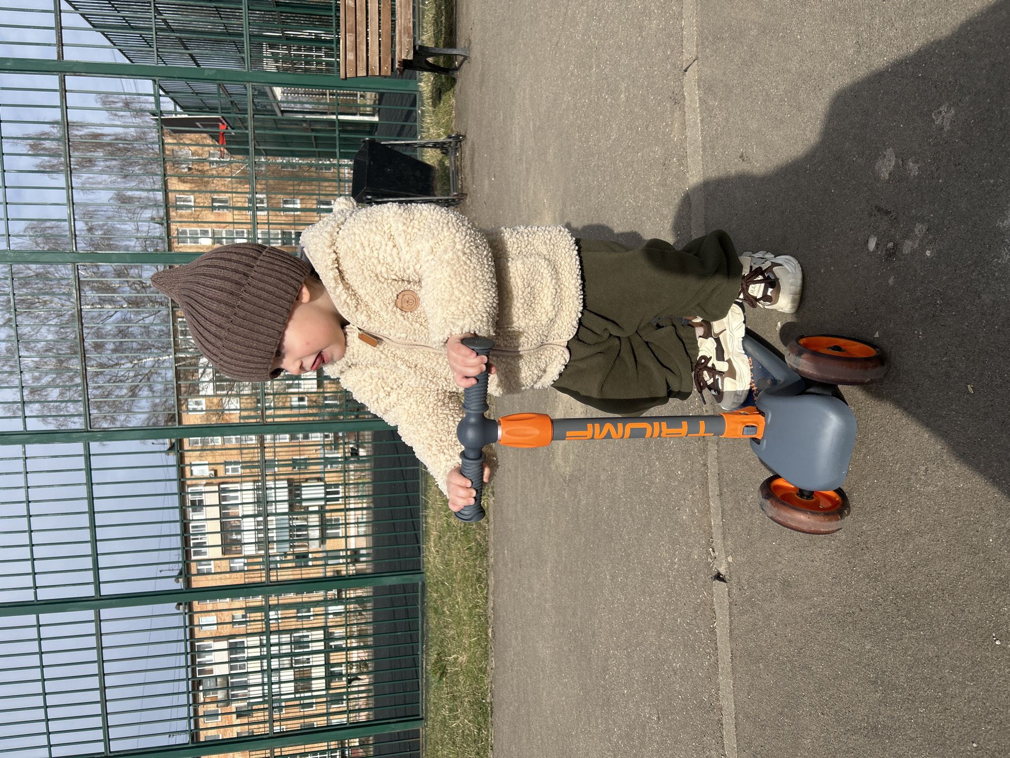 A child wearing a brown knitted hat, a cream-colored fleece jacket, and olive green pants stands on a gray tricycle scooter outdoors. The child is holding the handlebars of the scooter, which has an orange handle. The child's shoes are a combination of white, brown, and black. The background includes a green metal fence, a building reflecting in the fence, a gray paved surface, and a small patch of grass. Based on prior data and images, this photo was taken in Klin, Russia.