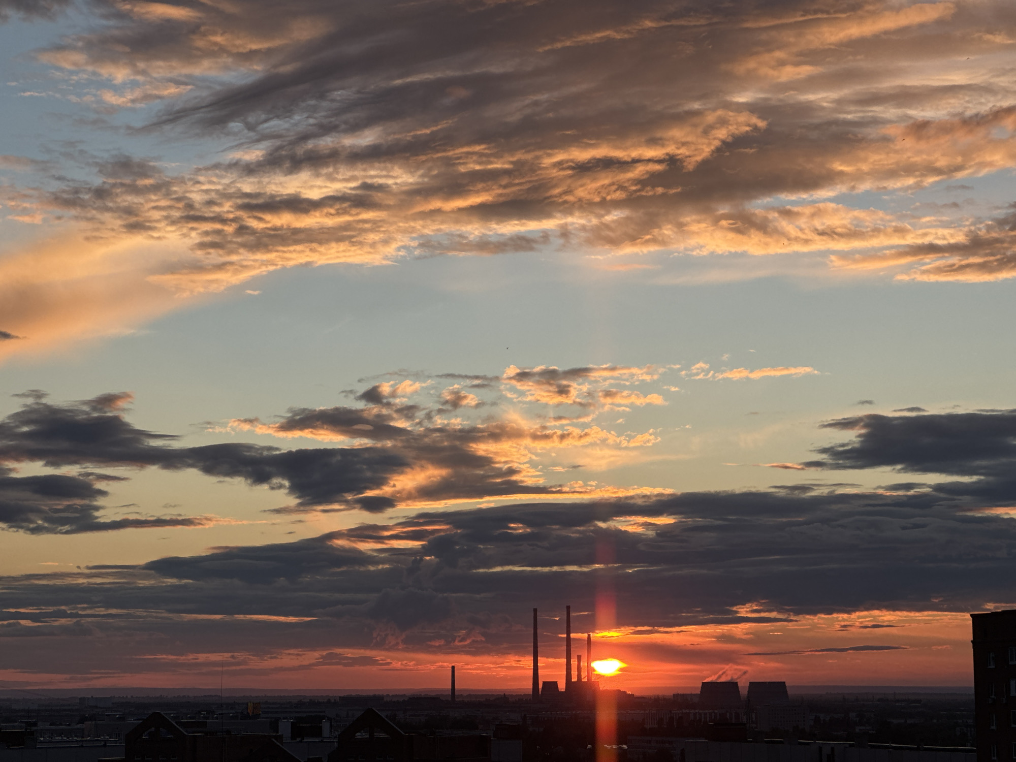 This is a photograph of a sunset over a city. The sky is filled with dramatic clouds, tinged with orange, pink, and gray. The sun is low on the horizon, partially obscured by clouds, and its rays are visible as beams of light. In the foreground, there is a cityscape with buildings and industrial structures, including several tall smokestacks from what appears to be a power plant. The overall impression is one of a vibrant and somewhat industrial urban landscape bathed in the warm glow of the setting sun.