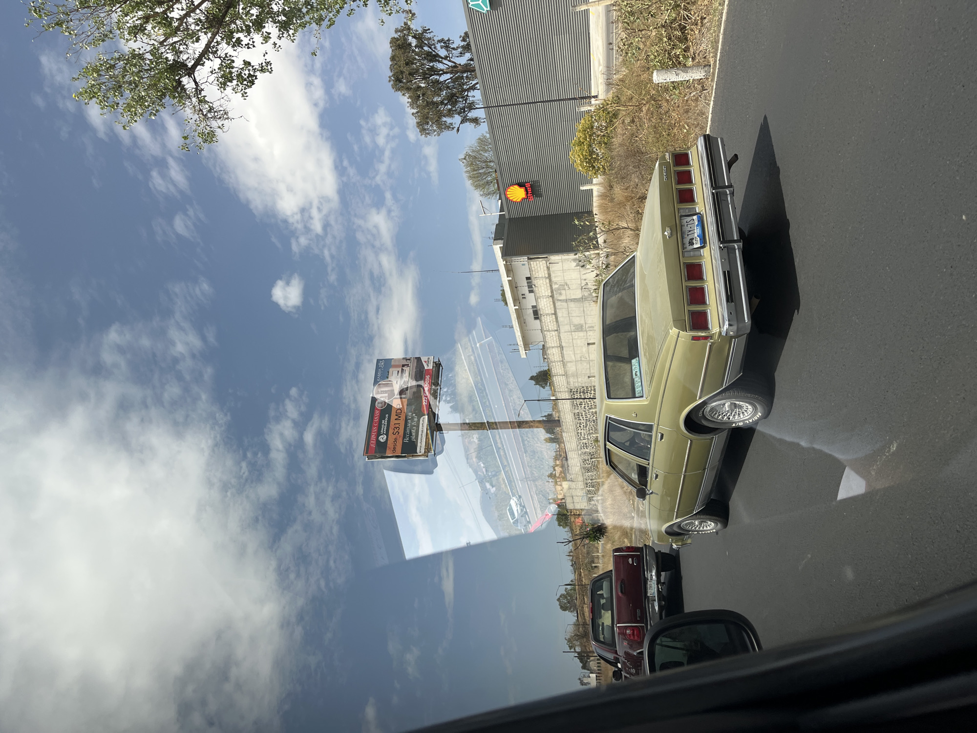 The image, taken from inside a vehicle, shows a traffic scene in San Bernardino Tlaxcalancingo, Mexico. The reflection on the car window includes a billboard advertising "JULIIMAS CASAS," a Shell gas station, and a partly cloudy sky. On the road, a vintage gold car is visible, followed by a maroon pickup truck. Other vehicles are queued in the traffic line. The overall environment is a mix of urban and roadside elements.
