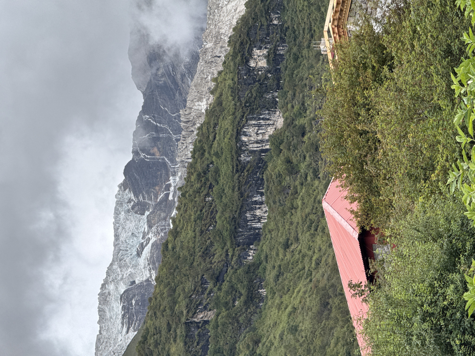 The image depicts a mountainous landscape in Samagaon, Nepal, under a cloudy sky. A large, snow-capped glacier dominates the upper portion of the frame, with meltwater streams cascading down the rocky slopes. The middle ground is covered in dense green vegetation, with darker rock formations visible beneath the foliage. In the foreground, a red-roofed building, possibly a house or guesthouse, is partially obscured by lush green trees and bushes. To the right of the red building, a portion of another structure with a stone base and decorative wooden elements is visible. The scene conveys a sense of remoteness and natural beauty, typical of a high-altitude village in the Himalayas.