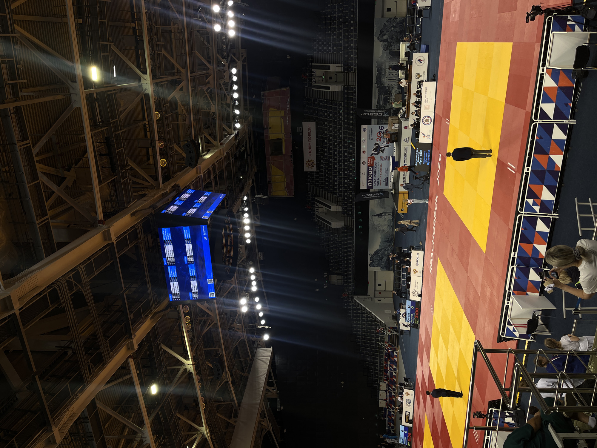 This media file depicts an indoor judo competition taking place in a large arena, identified as the "Krasnoyarsk League" in Russia. The central feature is a red and yellow judo mat, where an individual in a dark uniform, likely a referee or official, stands. Another person is visible near the mat's edge, and several people are seated at tables surrounding the competition area, possibly officials or coaches. One person is actively filming the event with a smartphone from the sidelines.
The arena has a high, industrial-style ceiling with visible metal trussing and numerous bright spotlights that illuminate the mat and surrounding areas. Large blue digital screens are suspended from the ceiling, displaying scores and information such as "Кра-ск5" (Kras-sk5) and "2:00" for a timer or score. One screen also shows "Дзюдо 05-06," indicating the event type and potentially a date or category.
Banners and signs around the arena provide further details. A prominent banner reads "КРАСНОЯРСКАЯ ЛИГА - ПАМЯТИ С.Н. БАЖЕНОВА 2006" (Krasnoyarsk League - In Memory of S.N. Bazhenov 2006), indicating the event's name. Another large banner displays "С ДНЁМ ПОБЕДЫ!!!" (Happy Victory Day!!!) alongside a sun logo, suggesting the event's timing or commemorative nature. Sponsor logos such as "МЕНТАЛПЛЮС" (MENTALPLUS), "СЕВЕР" (SEVER), and "JUDO.RU" are also visible. The barriers around the mat feature a design incorporating the colors of the Russian flag (blue, white, and red). Rows of empty seating are visible in the background, suggesting that while an event is ongoing, spectator presence might be limited. The setting is clearly a modern sports facility designed for such competitions.