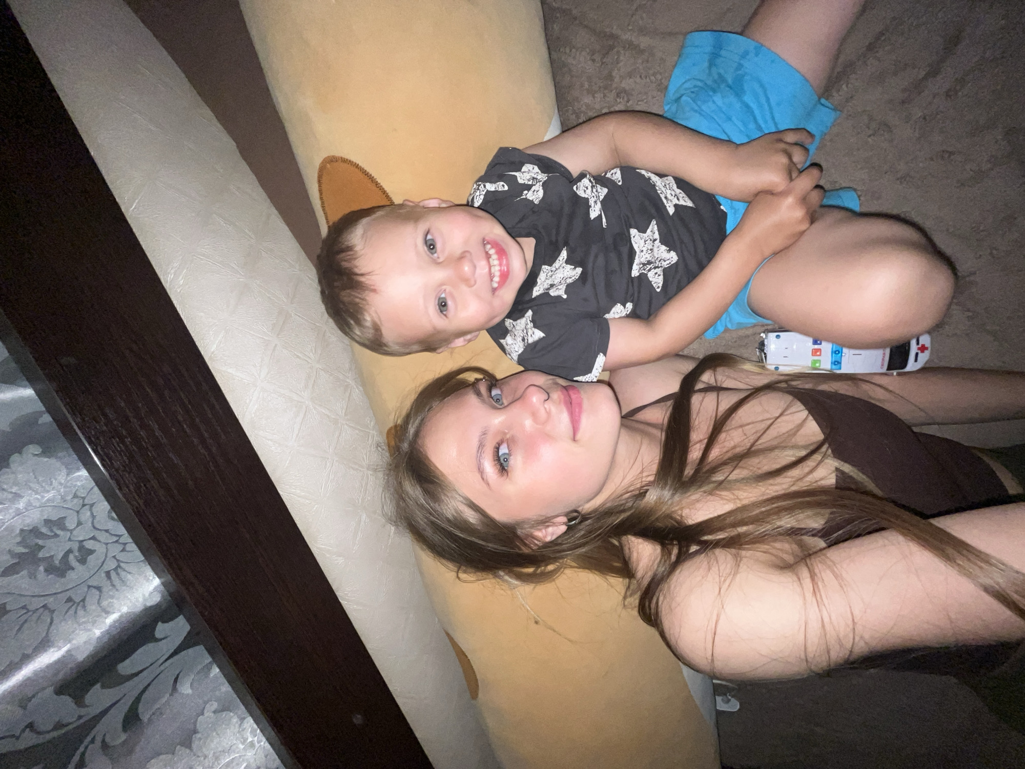 The image shows two people, a young boy and a young woman, lying down side-by-side on a couch or bed. The boy is in the foreground, looking directly at the camera with a wide smile. He is wearing a black t-shirt with white star patterns and blue shorts. The woman is lying behind him, her head resting on the same surface. She has long, brown hair and is looking upwards and slightly to her right. She is wearing a dark-colored tank top. A toy ambulance is visible between the boy's legs. The background appears to be a dimly lit room, possibly a living room or bedroom. The scene evokes a feeling of casual relaxation and familial affection. The lighting suggests it might be evening or nighttime. There is no visible text in the image. The location context is Фоки, Russia.