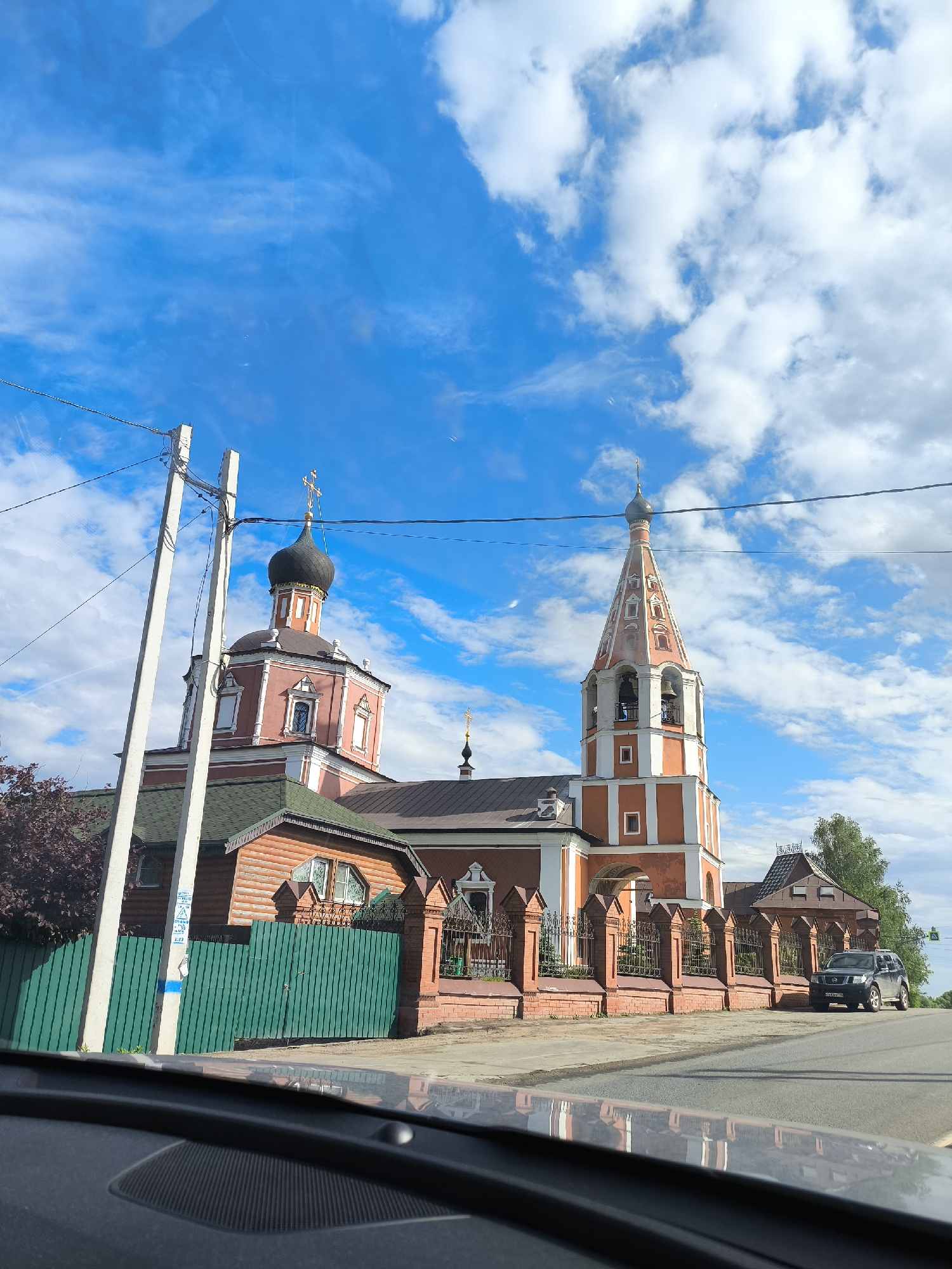 A street-level view from inside a vehicle presents a picturesque scene in the city of Unknown, Unknown. The architecture is dominated by a church complex featuring two prominent structures: one with a pink hue and a black onion dome topped with a golden cross, and the other a tall, orange-and-white bell tower, also topped with a cross.
A lower, wooden structure with a green roof connects to the church buildings. A green wooden fence stands in front of the wooden building, while a brick wall with wrought-iron fencing fronts the rest of the church property.
A black SUV is parked along the roadside. Utility poles and wires are visible in the background against a vibrant blue sky filled with puffy white clouds. The scene is viewed from the interior of a vehicle, with the dashboard visible in the foreground.