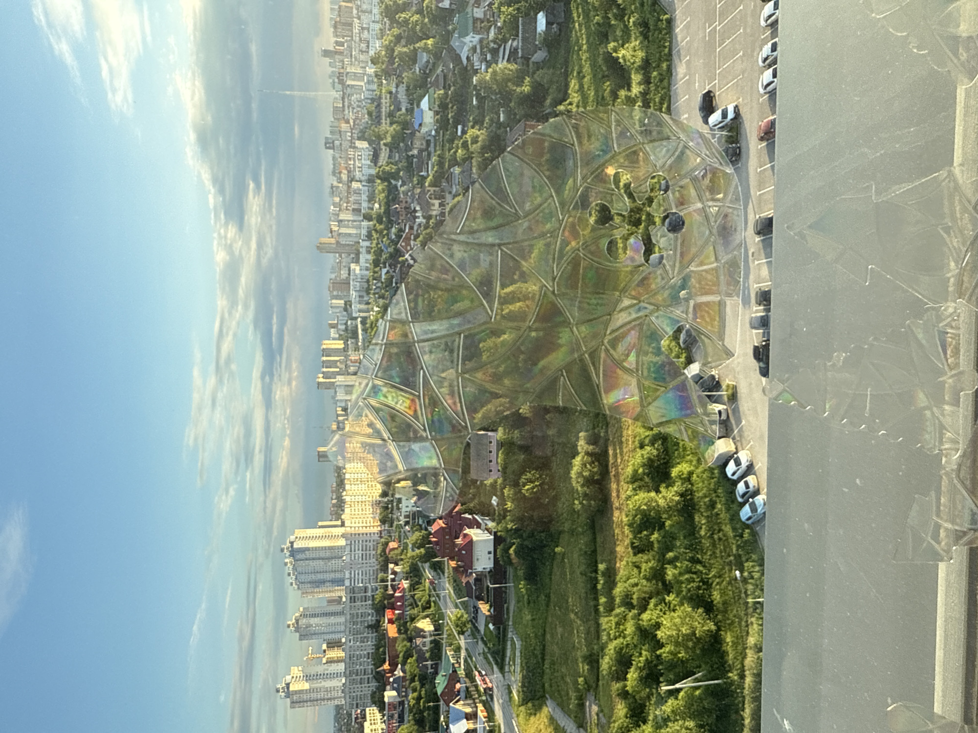 This media file shows an aerial view of a city and a park with a unique geodesic dome structure. The scene is set in Perm, Russia. The sky is blue with scattered white clouds, indicating fair weather during the daytime.
The foreground features a partially visible, modern glass geodesic dome with a colorful, iridescent finish. This dome appears to be a prominent object in the park or an open space. Beyond the dome, a densely populated urban area with numerous high-rise apartment buildings is visible. Adjacent to the dome and the urban sprawl is a lush green park area with trees and grass, interspersed with some houses. A parking lot with several cars is also visible near the dome.
The overall impression is one of a city bordering a green space, with a striking architectural feature. The time of day appears to be late afternoon or early evening, judging by the lighting and the long shadows cast by the buildings. There is no visible text in the image. The scene evokes a sense of urban planning and the integration of modern art installations within a natural or semi-natural environment.