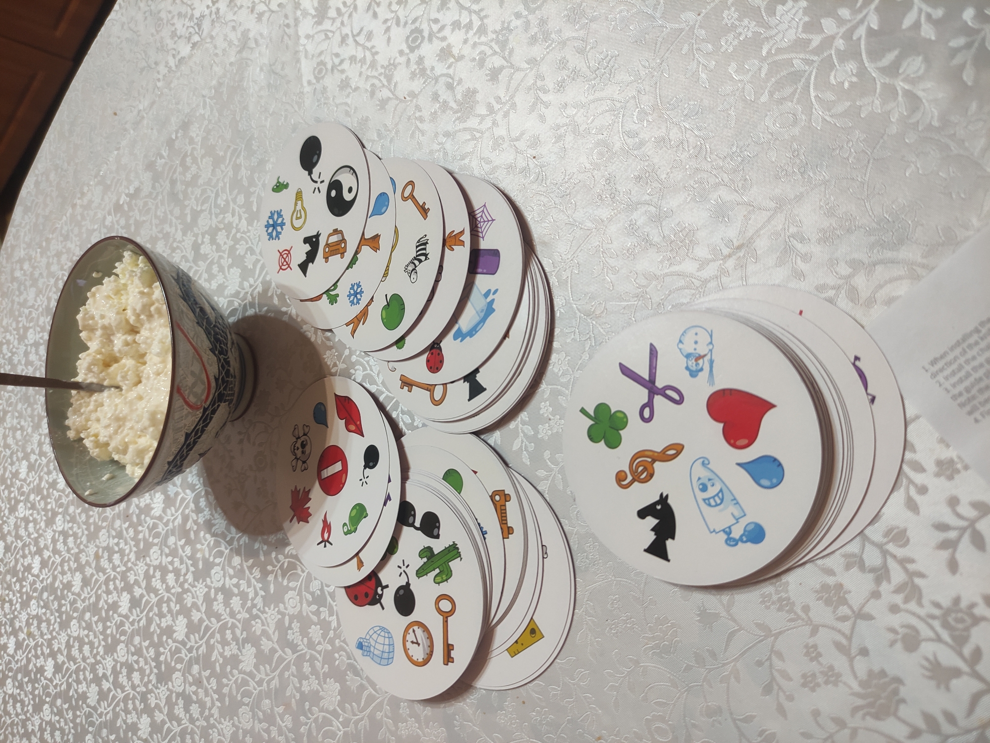 A bowl of cottage cheese sits on a white tablecloth next to two stacks of playing cards. The round cards contain various images, including a yin-yang symbol, a car, a key, and a ladybug. The scene is lit by indoor lighting. The location is Kratovo, Russia.