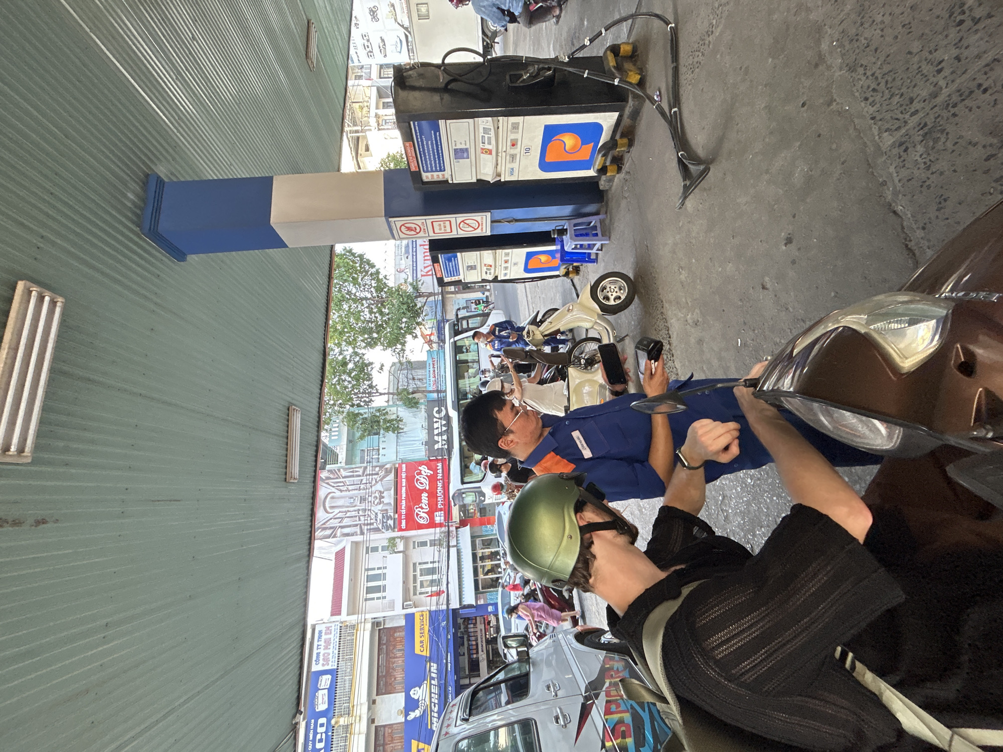 The image depicts a scene at a gas station in Tay Nha Trang Ward, Vietnam. In the foreground, a person wearing a black short-sleeved shirt and a green helmet is seen from behind, leaning over a brown motorcycle. Next to them, a gas station attendant in a blue uniform with orange trim is holding a device, possibly for payment or transaction processing. The attendant's badge is visible. In the background, a row of gas pumps with digital displays are visible, with hoses connected to them. Several motorcycles and a silver van are parked in the vicinity. Behind the gas station, buildings with signs in Vietnamese are visible, suggesting an urban setting. The overall environment appears to be an open-air gas station with a corrugated metal roof supported by a blue and white pillar. The lighting suggests it is daytime.