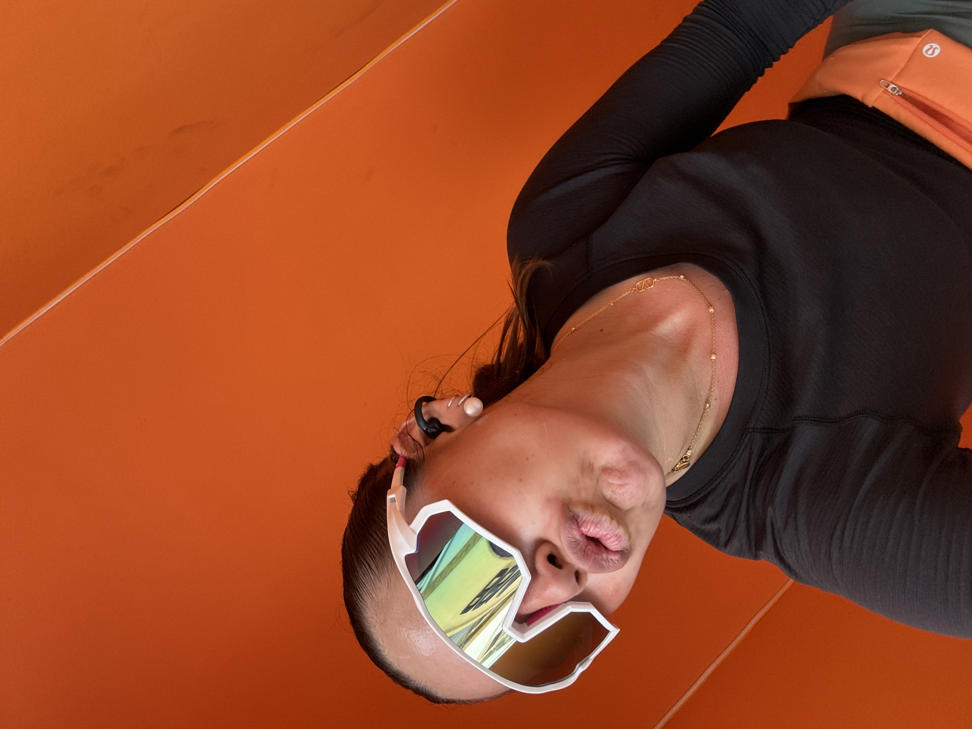 A woman is posing with a pouting face, wearing white-framed sunglasses with iridescent lenses and a black, long-sleeved shirt. She has an orange waist bag and a gold necklace. The background is orange, suggesting an urban setting, possibly in Puebla, Mexico, given its known vibrant and colorful architecture. The composition appears intentionally tilted, giving a disorienting perspective.