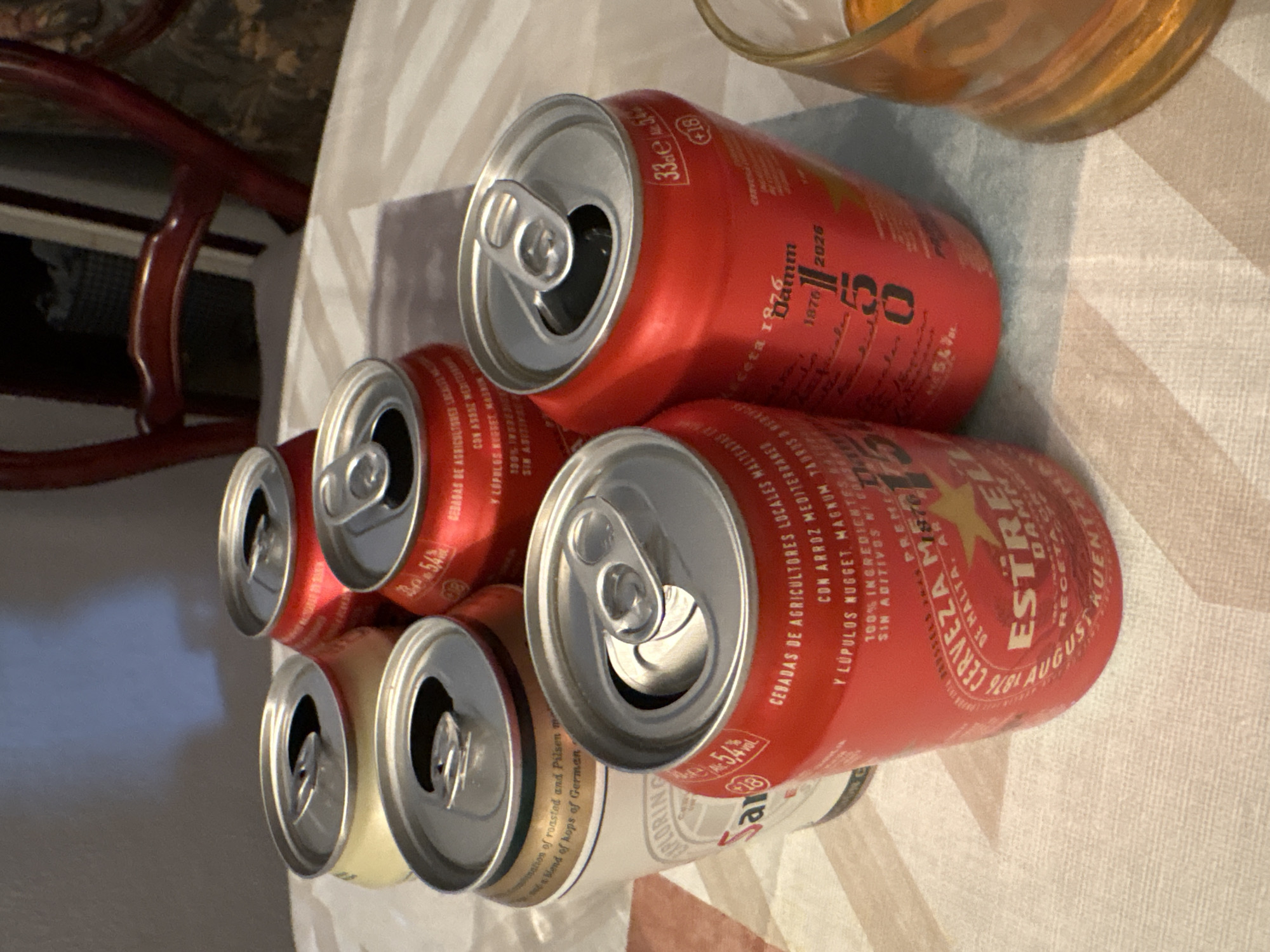 The image shows a close-up of six open beer cans on a table covered with a striped tablecloth. There are four red cans of Estrella Damm beer and two white and gold cans of an unidentified beer. The Damm cans are partially covered in text, advertising the 150th anniversary of the company and stating "Cerveza de Malta". There is a glass with a yellowish liquid on the top right. A dark brown chair is visible on the left. It seems to be a casual setting.