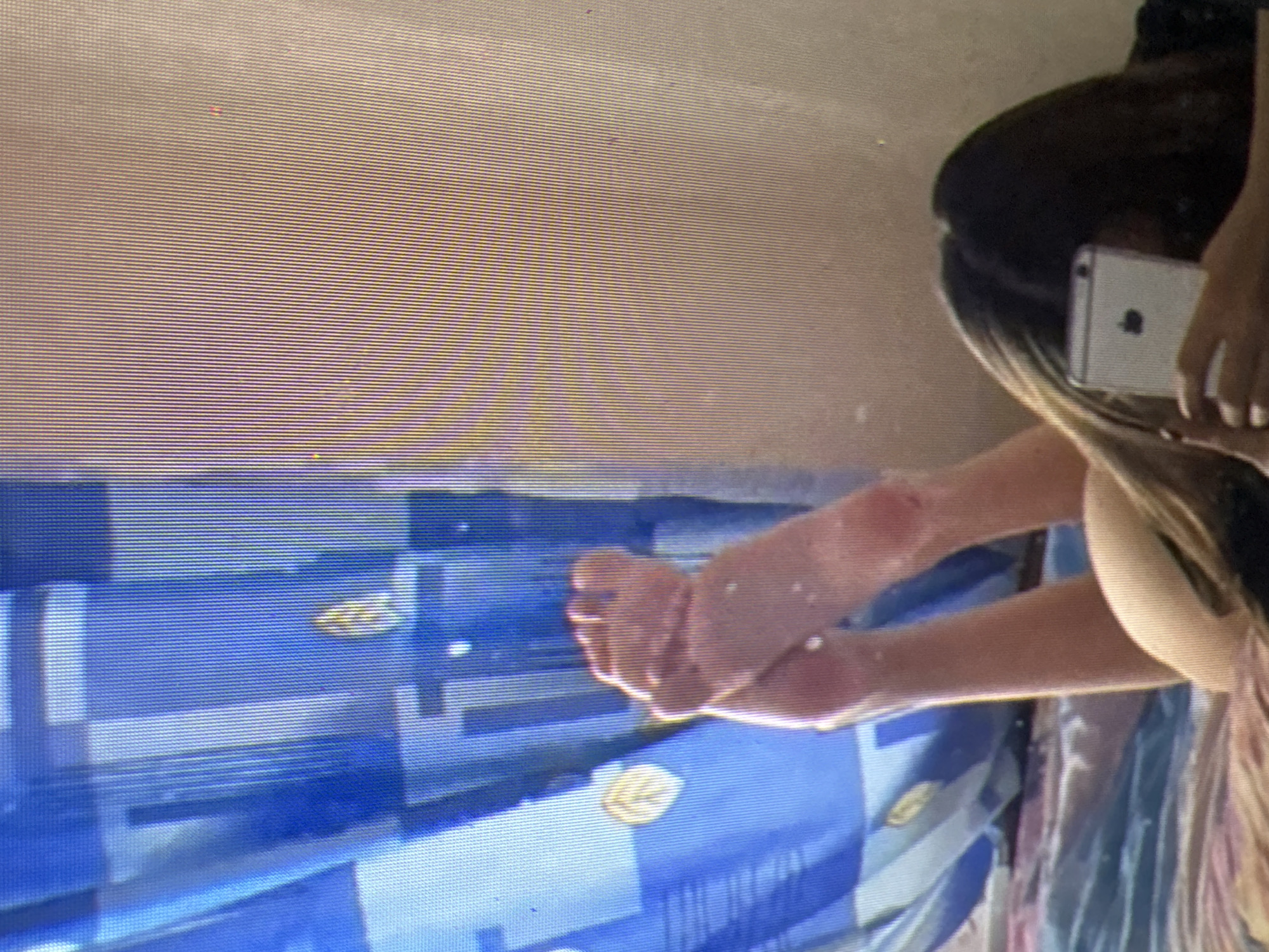 In a bedroom in Castelo Branco, Portugal, a person is lying on a bed, taking a selfie with their phone. The person's feet are visible in the foreground, propped up and pointing towards the camera. They are wearing a white t-shirt, and their dark hair is spread out around them. The background is cluttered with a blue and white striped curtain and a wall. The overall impression is one of casual intimacy and self-documentation.