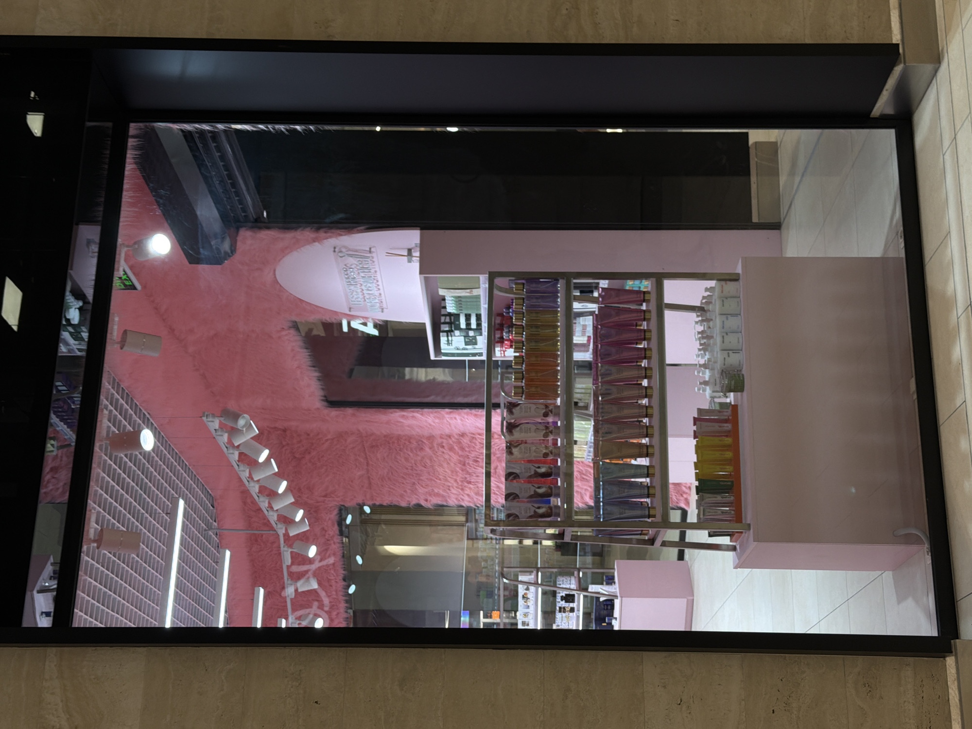 This is a photograph of a retail store window display, likely in a shopping mall. The storefront is decorated with bright pink, fluffy material on the walls and ceiling. Various beauty products are displayed on shelves, including rows of bottles and tubes. In the foreground, a metal shelf unit holds numerous cosmetic products. Behind this, a pink display case is visible. The store's interior appears to be brightly lit, with spotlights on the ceiling. The reflection on the glass indicates the presence of overhead lighting and the surrounding mall environment. There are no people visible in the image. The overall aesthetic is feminine and commercial, emphasizing brightly colored products in a clean, well-lit setting. The specific location in Kyiv, Ukraine, cannot be determined from the image alone.