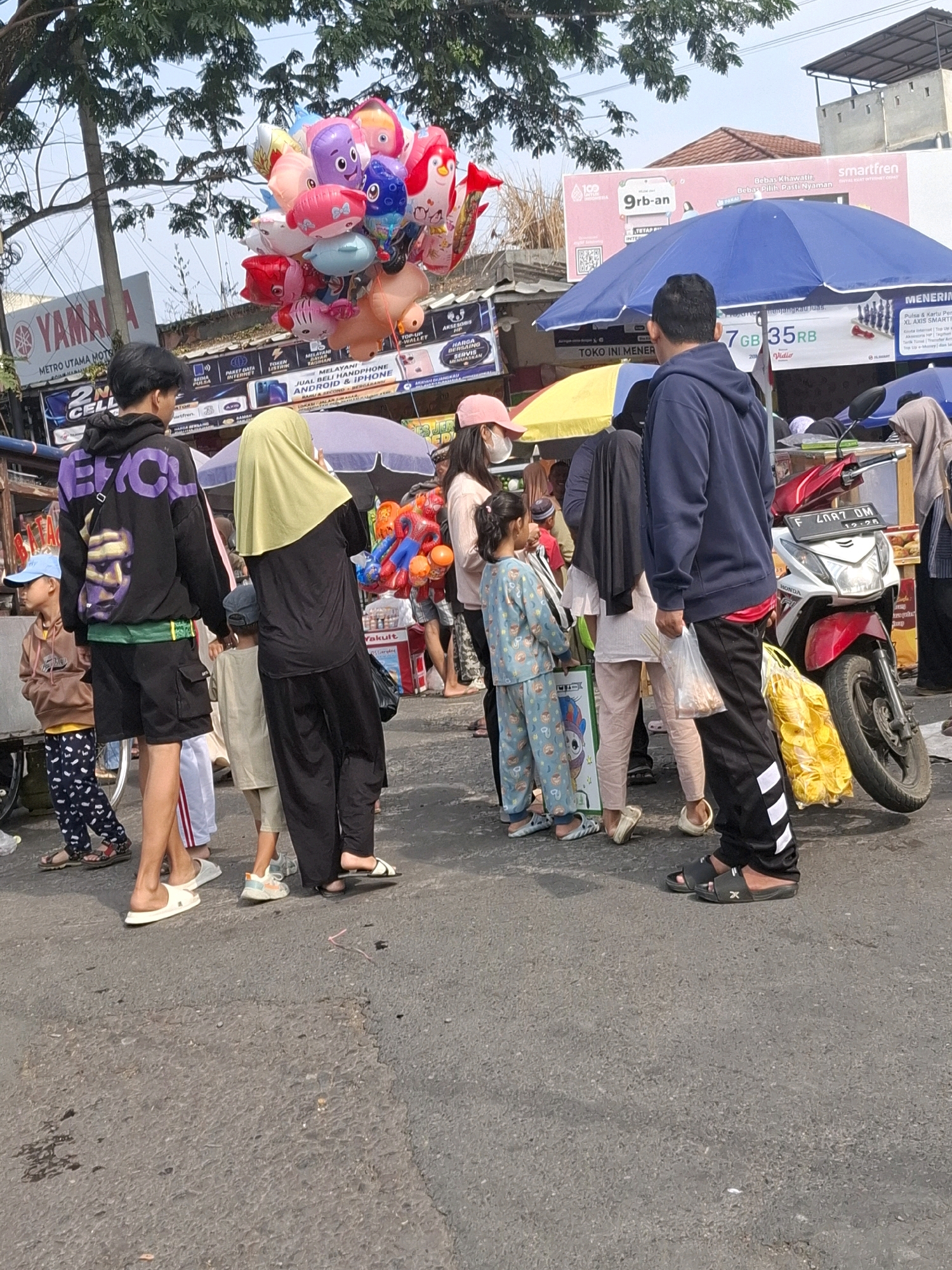 This image depicts an outdoor scene in a marketplace in Karangtengah, Indonesia, likely on a street with vendors. In the foreground, a group of people are gathered, some looking at merchandise and others walking. A man in a dark blue hoodie and black pants with white stripes is standing near a red and white motorcycle. To his left, a woman in a light green hijab and black clothing is holding a string of colorful balloons, which are prominent in the upper left quadrant of the image. A young girl in a light blue patterned outfit is standing near the balloons, facing away from the camera. Another young boy in a brown hoodie and patterned pants is visible on the far left, looking towards the scene. Various market stalls with umbrellas are set up in the background, displaying merchandise and advertisements. The overall environment suggests a busy, casual marketplace.