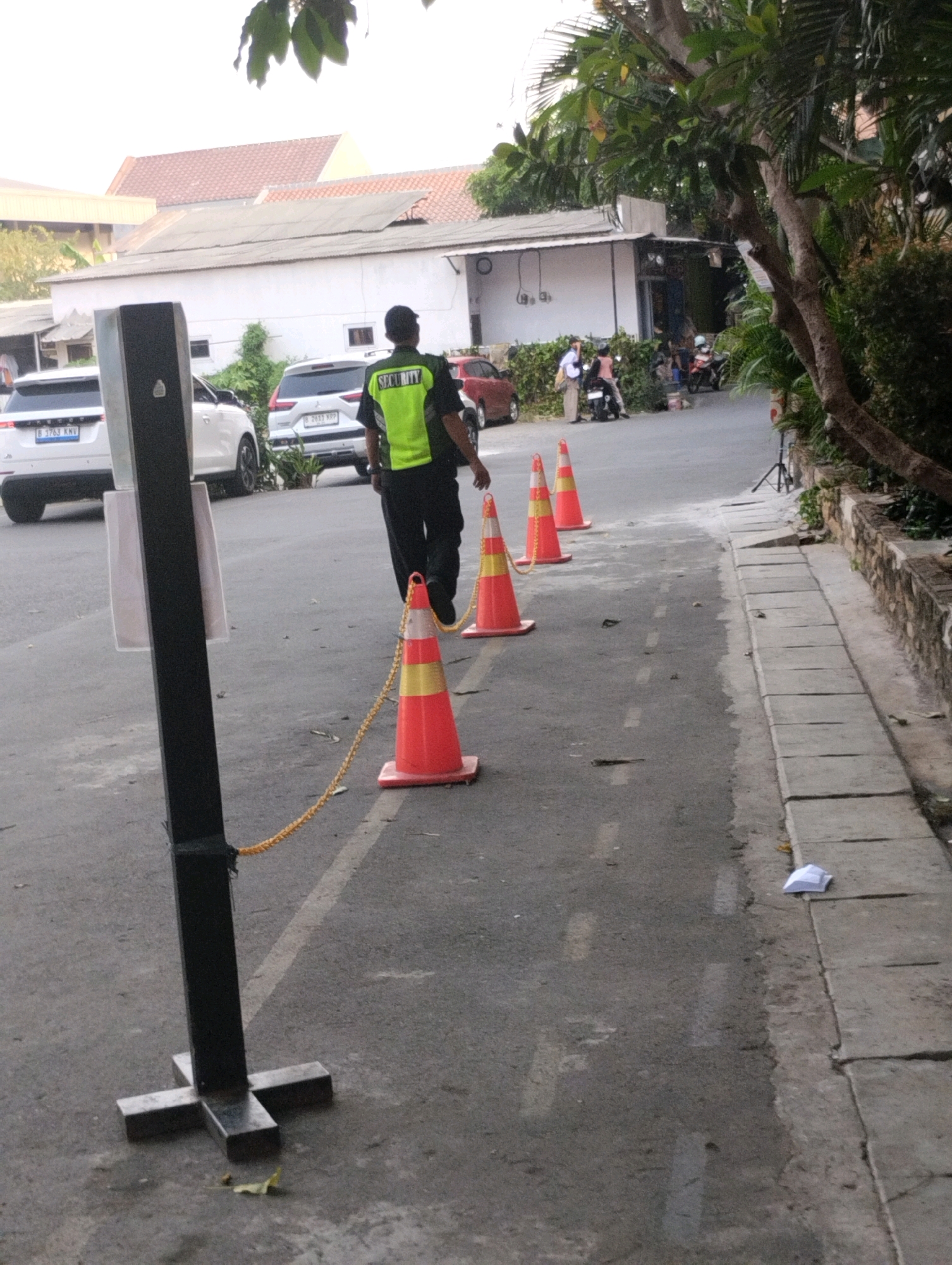 The image shows a street scene in Bekasi, Indonesia. In the foreground, a black metal stanchion with a cross-shaped base stands on the asphalt. A yellow rope is attached to the stanchion and stretches towards the right, connecting to a line of orange traffic cones with yellow stripes. A man in a bright yellow vest with "SECURITY" printed on it walks away from the camera, towards the right, past the cones. Behind him, several white cars are parked on the street. A white SUV with license plate B 3763 KNV is on the left, and another white SUV with license plate B 2631 XPP is further down the road. To the right of the street, a sidewalk with concrete slabs runs alongside a stone wall, with lush green vegetation behind it. Buildings with white walls and reddish-brown roofs are visible in the background. The sky is overcast, suggesting it may be daytime with diffused light.