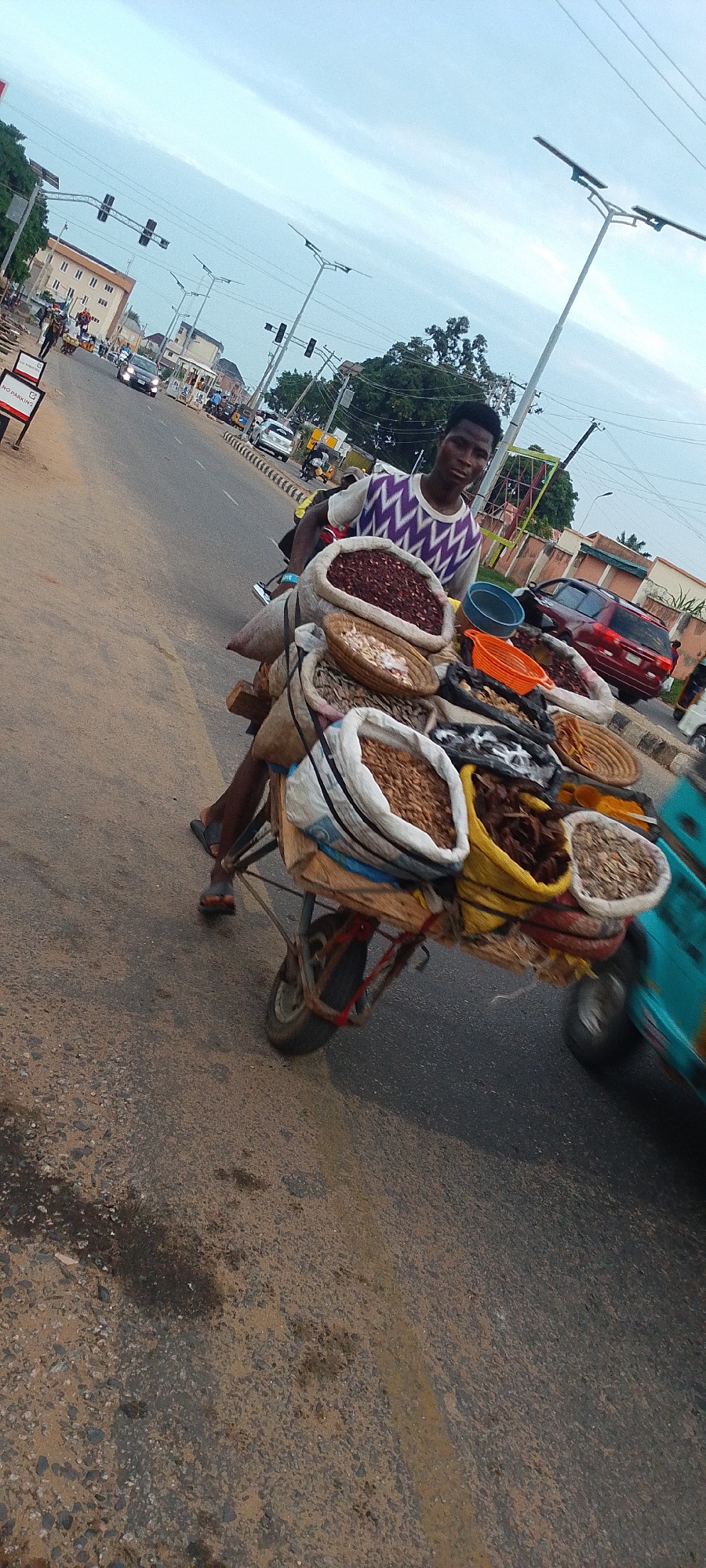 In Lokoja, Nigeria, a young man sits on a tricycle, laden with bags and baskets of foodstuffs, likely for sale. He wears a light-colored shirt with a dark zigzag pattern on the chest. The tricycle is positioned on a paved road with moderate traffic, including cars, motorcycles, and another tricycle. The environment shows a typical urban setting with buildings, traffic lights, and streetlights.