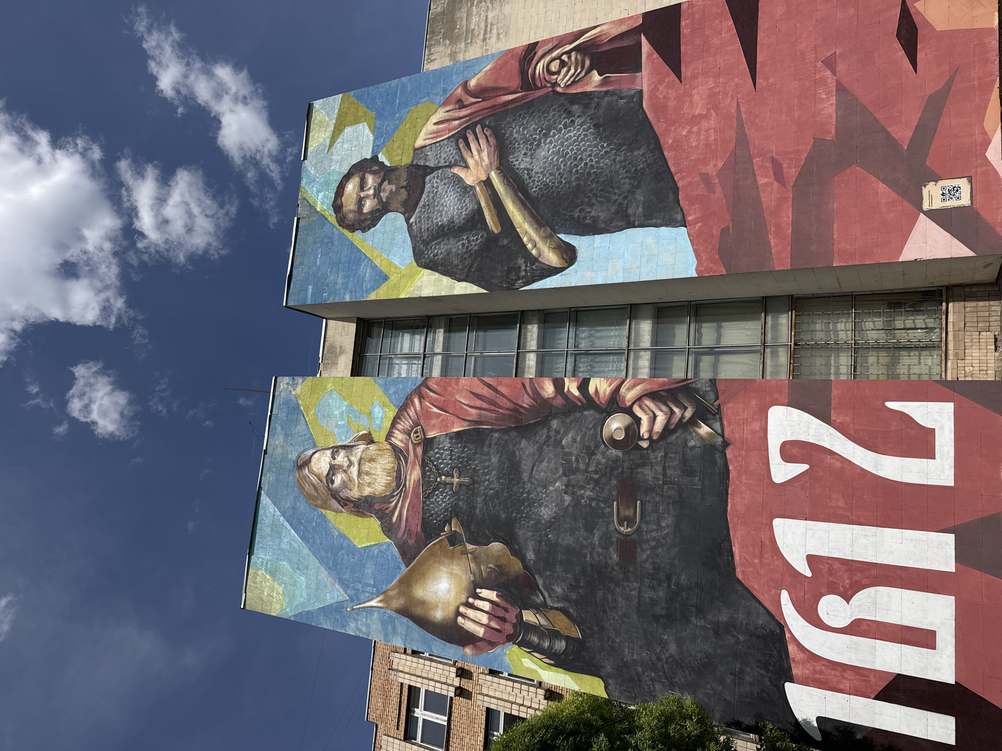 A low-angle shot of a building in Unknown, Unknown, displaying two large mural paintings. The top mural depicts a man with short, dark hair and a beard, wearing dark armor and a red cloak, set against a blue and yellow background. The lower mural features a man with a long, light-colored beard, also wearing dark armor and a red cloak. He is holding a golden object. The background is also blue and yellow, and the large, white number "1812" is painted on the right side of the mural, overlapping the red background. The sky above the building is blue with scattered white clouds. A portion of a brick building is visible at the bottom left, with a green tree obscuring part of it.