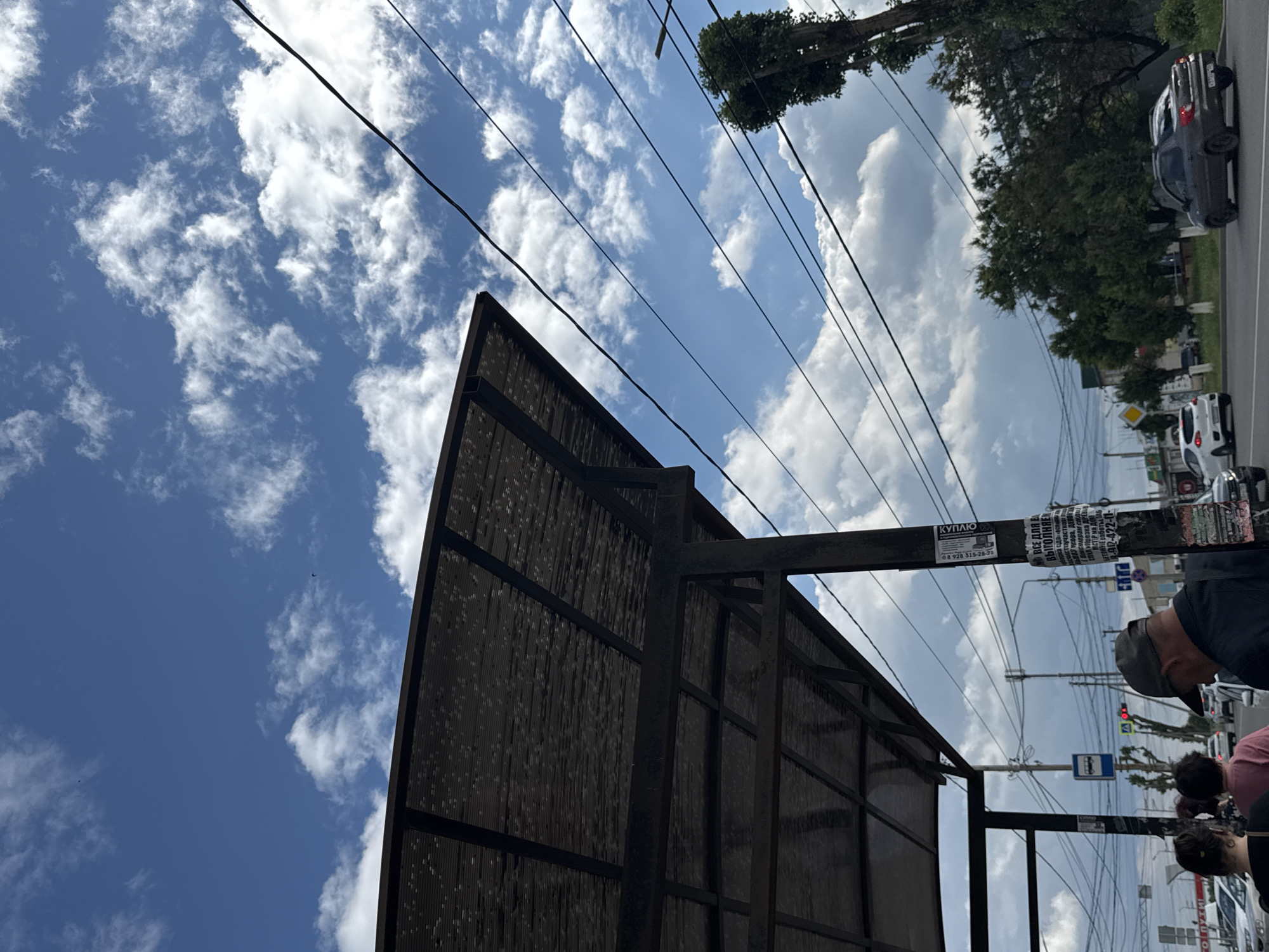 In Mikhaylovsk, Russia, the image captures a street scene from a low angle, looking upwards towards a cloudy sky with patches of blue. The perspective is slightly tilted. Prominently featured is the dark, angular structure of a billboard or a similar outdoor advertising panel. This structure has a metal frame and a dark, textured surface. A street lamp post is attached to this structure, adorned with various flyers and advertisements. One flyer reads "КУПЛЮ" (I will buy) and a phone number "8 928 315-28-28". Another poster has text in Cyrillic, including phrases like "ВСЕ ДЛЯ ВА ВЫГОЛНЯЕМ" (We do everything for you) and mentions services like "БЕТОННЫЕ работы" (concrete work).
In the background, a paved road is visible with cars driving on it, suggesting it's a public street. Trees line the roadside, their green foliage contrasting with the sky and the dark structure in the foreground. Power lines crisscross the sky, adding to the urban setting. Several people are visible near the bottom of the frame, appearing to be waiting or passing by, their faces partially obscured. One person wears a grey cap. The overall impression is of a typical daytime scene in a Russian town or city, with everyday activities and advertisements. The sky suggests it's a bright day, possibly with the possibility of rain due to the clouds.
