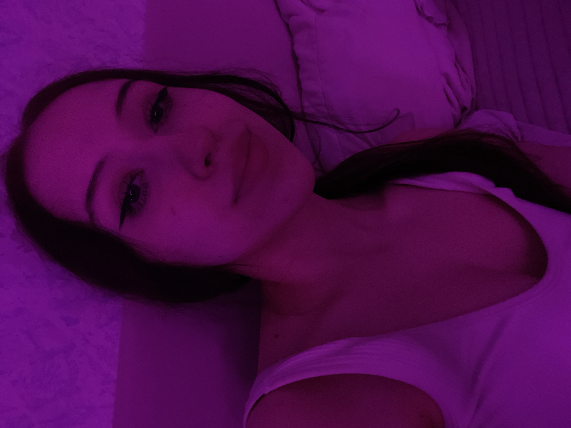 The image shows a young woman with dark hair, wearing a white tank top. She is looking towards the camera with a slight smile. The lighting in the image is predominantly purple, casting a strong hue over the scene. The background appears to be a textured surface, possibly a wall, and a pillow. The woman's hair is pulled back in a ponytail. Her makeup includes eyeliner and mascara. There is a mole visible on her neck. The scene is indoors, and the overall impression is of a casual, intimate setting. The image does not provide any information about the location in Votkinsk, Russia.