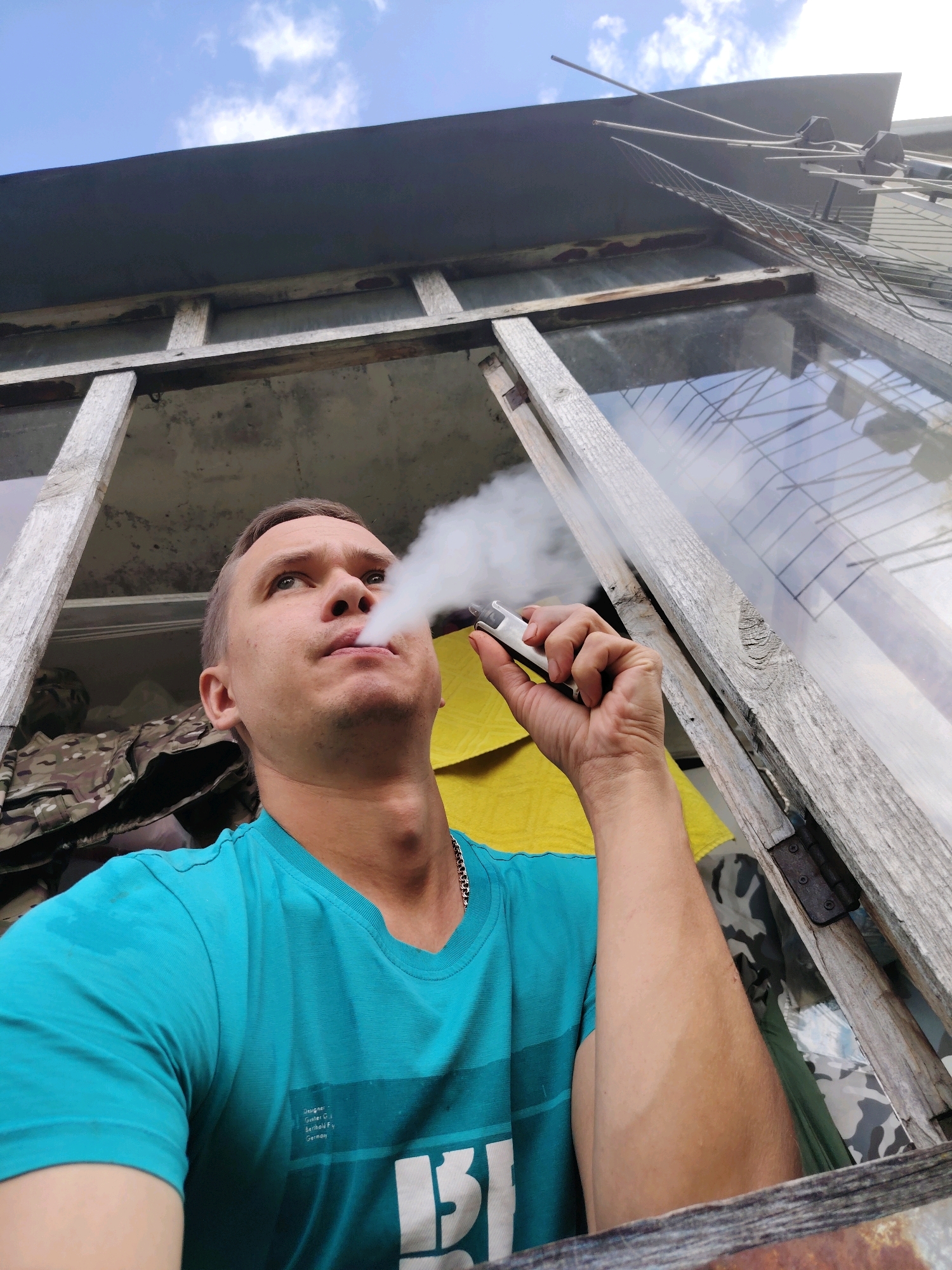 A low-angle, upward shot captures a man exhaling vapor from a vape pen. He is positioned in the lower half of the frame, looking upwards and to the left. He wears a teal t-shirt with a white graphic and a silver chain necklace. His left hand holds the vape pen, which is white and silver, emitting a plume of white vapor. The background consists of an open, dilapidated window frame with weathered wooden elements. Through the window, one can see a gray, possibly concrete, surface and a blue sky with scattered white clouds. To the left of the man, a camouflage-patterned fabric is visible. The image appears to have been taken outdoors, likely in a residential area or an abandoned structure.