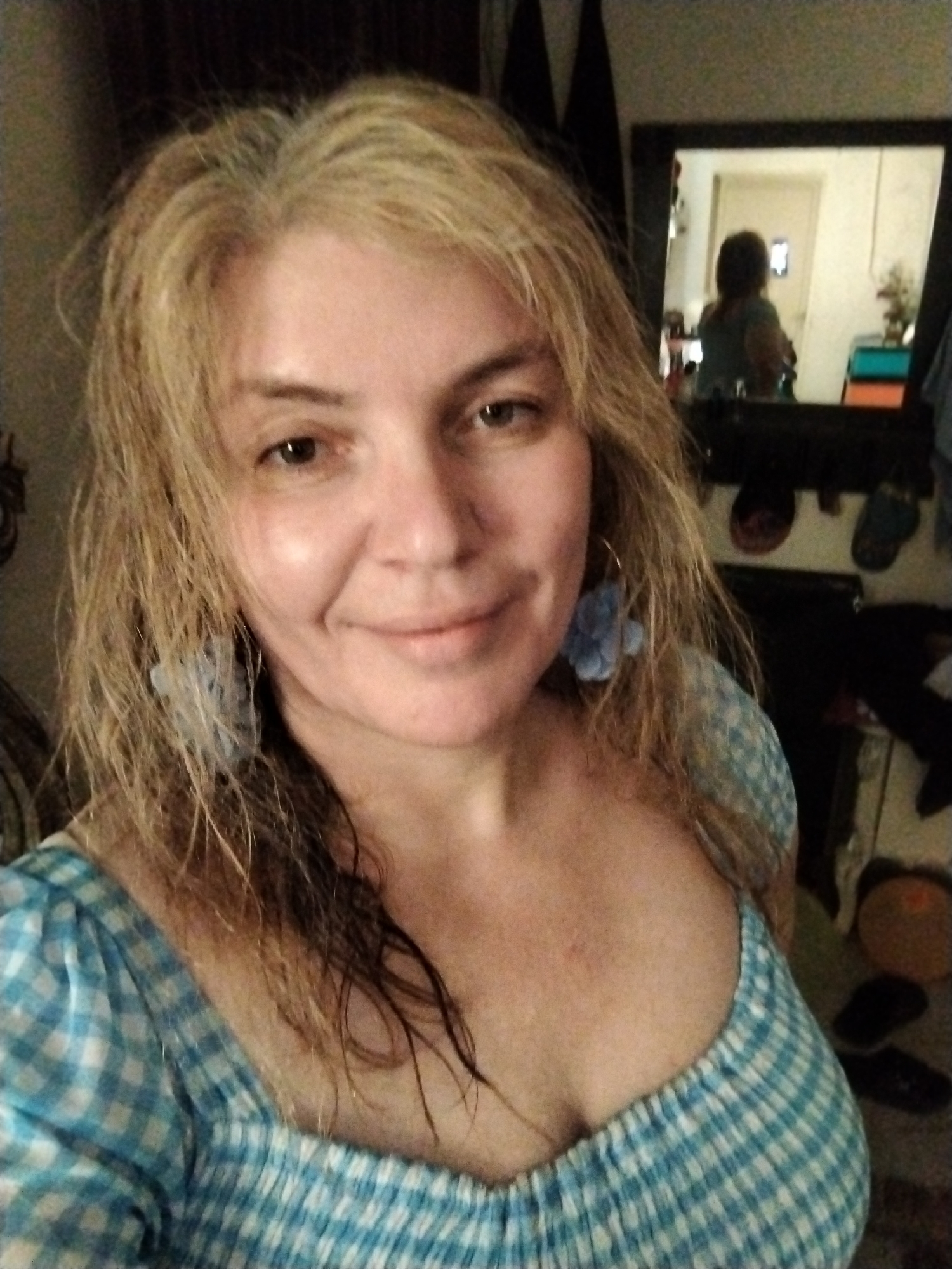 A woman is in a room, likely a bedroom, with a mirror visible in the background reflecting a portion of the room. The woman is facing the camera with a slight smile. She has blonde hair and is wearing a blue and white checkered dress. She also has blue flower earrings. The reflection in the mirror shows another person in the background. The location is Al Mas'oudi, United Arab Emirates. Other objects are visible in the room, including shoes near the mirror.