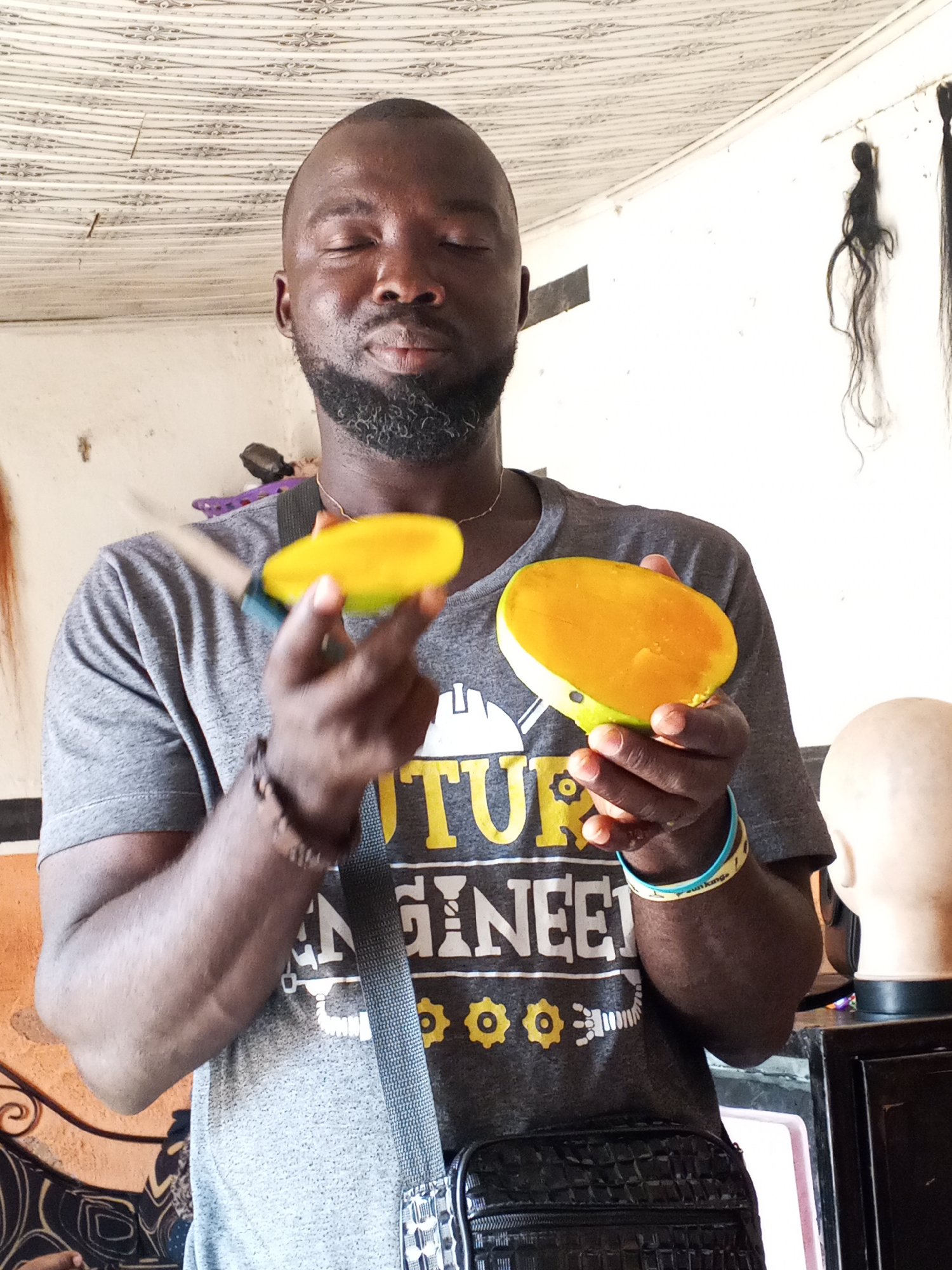 A man with dark skin, a shaved head, and a short, graying beard stands indoors holding two halves of a mango fruit. He is wearing a gray t-shirt with the words "FUTURE ENGINEER" and illustrations of gears and a building hard hat. A gray strap crosses his torso, attached to a black bag. He has a bracelet on his left wrist.
In the background, there is a white ceiling and a white wall. To the right, hanging hair extensions are visible. A mannequin head is also present on a dark surface. A knife is present in one of the mango pieces.
The location is identified as Gwong, Nigeria.