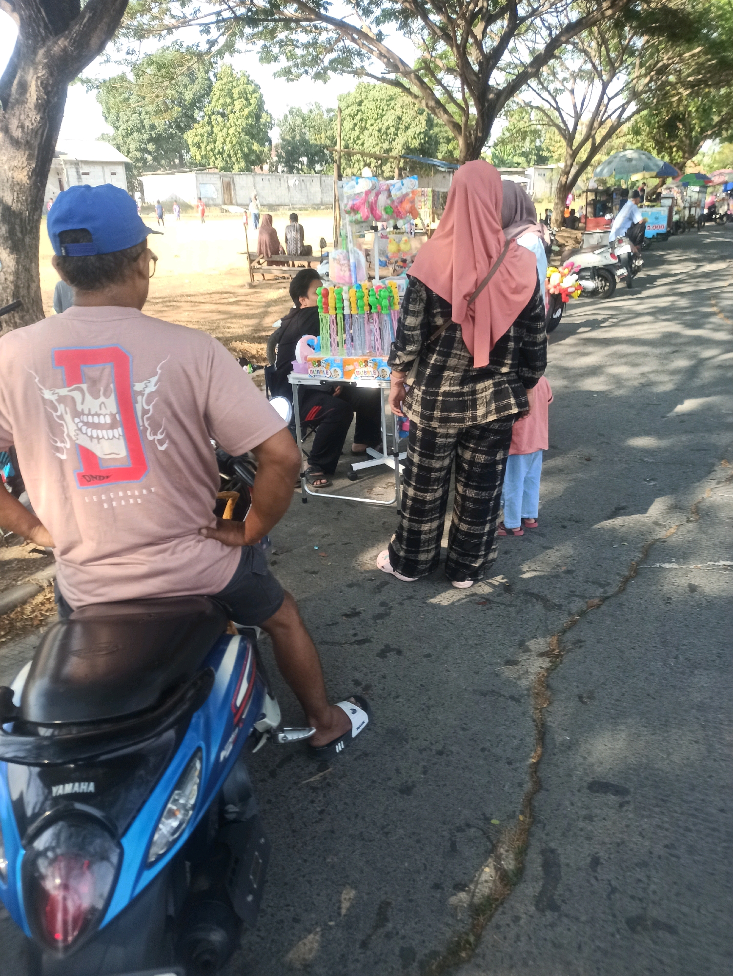 In Telagamurni, Indonesia, an outdoor scene unfolds on a paved road. The foreground features a man seated on a blue Yamaha motorcycle, his back to the camera. He wears a light brown t-shirt with a graphic design on the back and dark shorts. In the mid-ground, a woman in a plaid outfit and headscarf stands with her back to the viewer, facing a stall displaying various colorful toys and bubble wands. To her left, another woman in a pink headscarf is partially visible. A young boy is seated behind the stall. In the background, several other individuals are visible near a row of buildings, and more stalls line the roadside, some with umbrellas providing shade. Trees with dense foliage line the area. The atmosphere appears to be a busy marketplace or gathering.