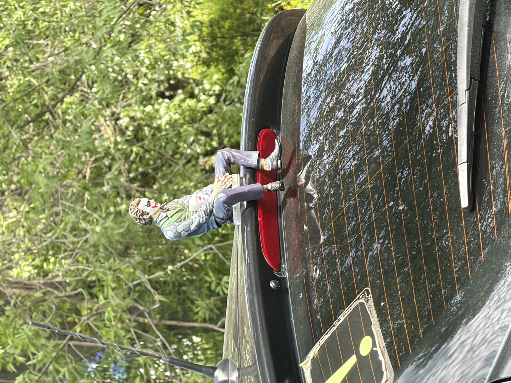 A darkly tinted car is parked outside in a green, leafy area. A figurine of the Joker is positioned, seemingly stuck, between the rear spoiler and the rear taillight. The Joker figurine is dressed in a green hexagon-patterned jacket, light-grey trousers, and trainers. It's difficult to discern its features due to lighting and distance. The back windscreen is dirty and has heater elements visible. The overall impression is one of quirky decoration on a vehicle. No information indicates this is in Izhevsk, Russia.