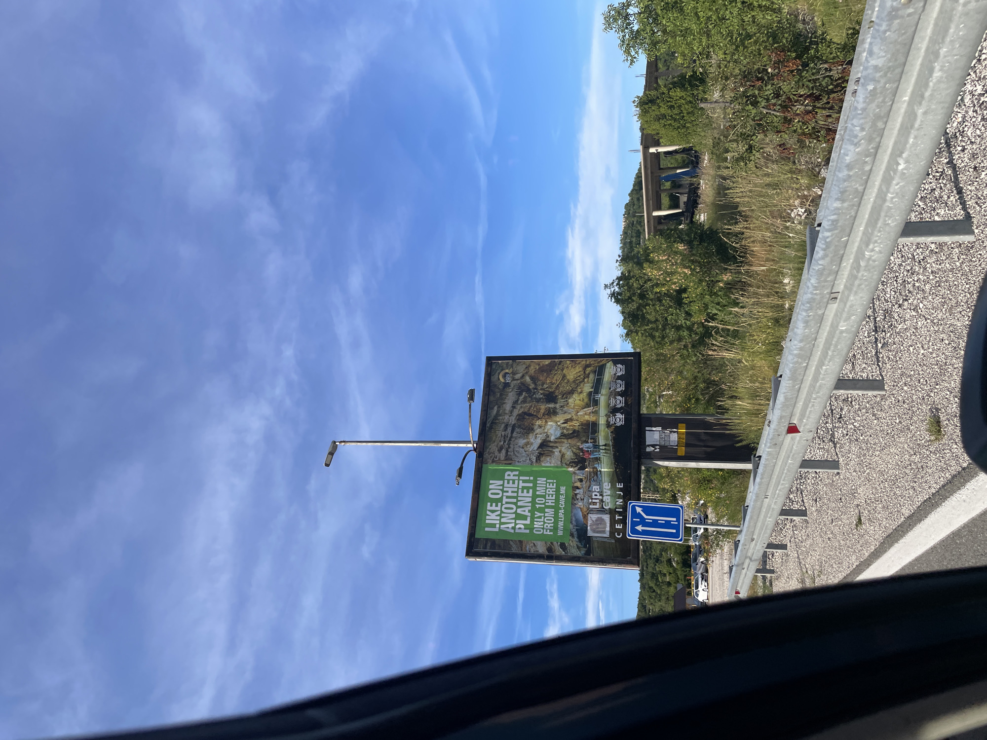 The image shows a roadside billboard in Očinići, Montenegro, advertising Lipa Cave. The billboard features an image of a cave interior with a group of people, and text that reads "LIKE ON ANOTHER PLANET! ONLY 10 MIN FROM HERE! WWW.LIPA-CAVE.ME" along with "Lipa Cave" and "CETINJE". Below the billboard, a blue road sign with white arrows indicates two lanes for going straight. A silver guardrail runs along the side of the road, which is covered in gravel. In the background, there are green trees and a partially constructed building. The sky is blue with wispy clouds.