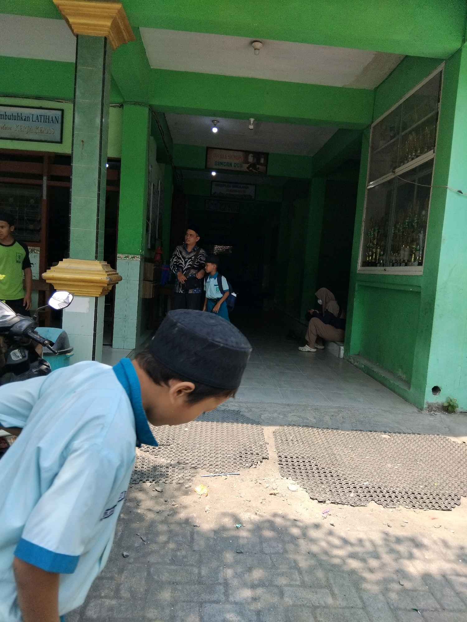The image captures a scene outside what appears to be a school or community building in Kepuh Kiriman, Indonesia. The building's facade is painted a vibrant green, with white trim around the ceiling. A prominent yellow pillar with decorative molding stands near the entrance.
In the foreground, a young person wearing a light blue shirt and a black kufi is facing away from the camera, seemingly looking down. A motorcycle is parked to the left of this individual, with a blue trash can visible behind it.
Further into the background, a person in a black and white patterned shirt and a kufi stands next to a younger person in a light blue uniform and a backpack, possibly a student. Another person, wearing a hijab and light-colored clothing, is seated on the ground in the deeper recess of the building's entrance.
Above and behind the seated individual, a sign with text in Indonesian, possibly referencing "DENGAAN DUA," is visible. To the right, a large window displays rows of trophies. The ground is a mix of paved surfaces and some dirt, with shadow patterns from overhead foliage. The overall environment suggests a sunny day.
