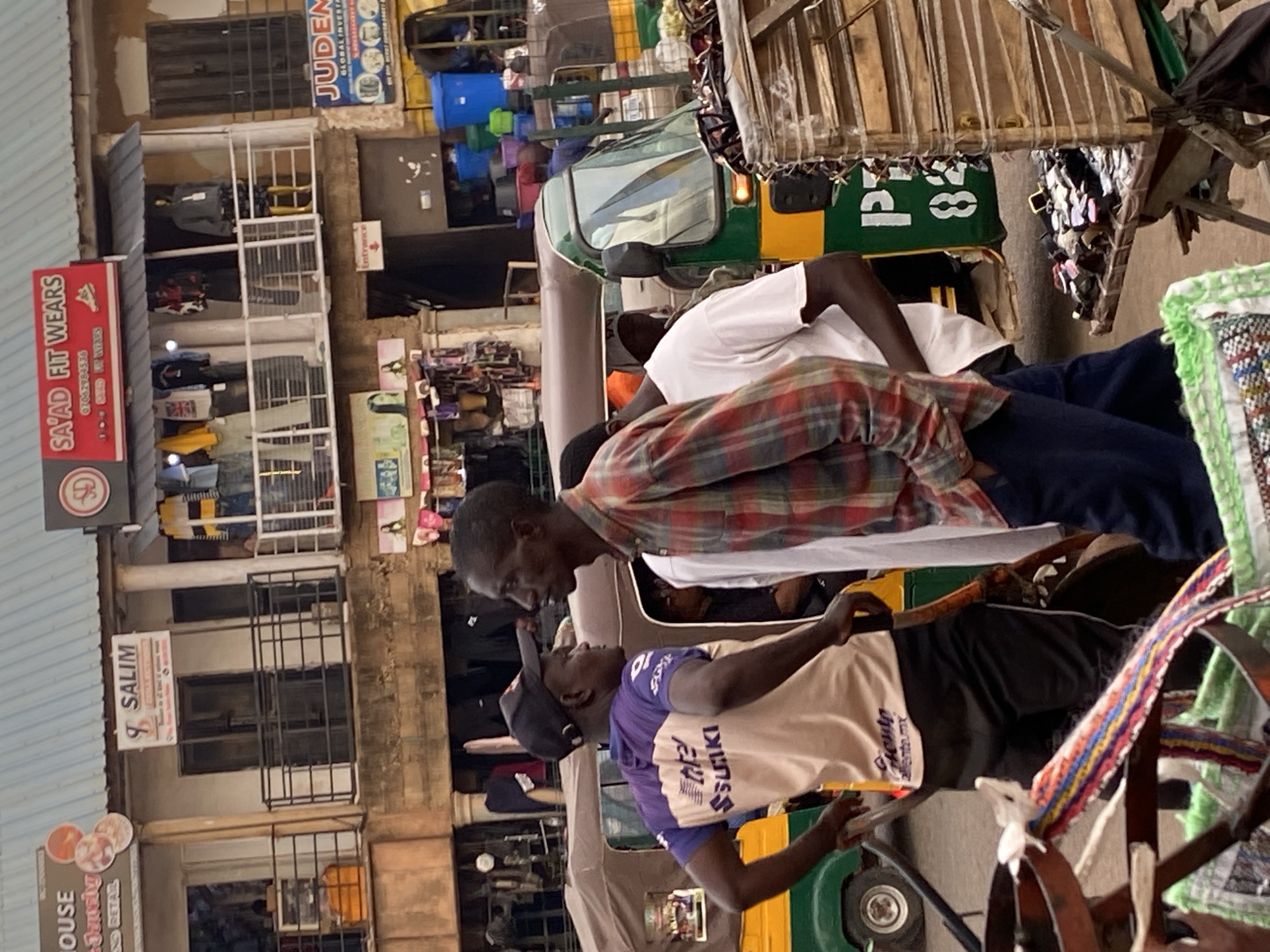 The image, rotated 90 degrees counter-clockwise, depicts a vibrant daytime street scene in Bukuru, Nigeria, characteristic of a bustling market area. In the foreground, two men are engaged in conversation. The man on the left wears a red, black, and white plaid shirt over a white undershirt, leaning slightly forward. Facing him, the man on the right wears a purple and beige t-shirt with "SSUAKI" printed on it, along with a black baseball cap. He appears to be seated or leaning within a green and yellow motorized rickshaw or tuktuk, which features a wooden canopy structure.
The background reveals a dense urban environment with multi-story buildings housing various shops and stalls. Open-front stores display clothing items, including one with a Union Jack t-shirt and another with yellow apparel. Stacked blue and green plastic containers are visible on a vehicle or stall further back, contributing to the busy market atmosphere.
Several signs are visible throughout the scene. A prominent red sign on the left displays "SA'AD FIT WEARS," along with a phone number "07062984536" and the slogan "GOOD SET WEARS." Below this, a white sign reads "SALIM." Higher up on a building, a blue sign advertises "JUDEN GLOBAL INVEST" with "allenta.mx" visible below. A smaller white sign with an arrow clearly indicates "Entrance." On the rear of the green and yellow vehicle, the number "P28" is visible. Near the bottom left, part of a sign reads "HOUSE RETAIL." The overall impression is one of active commerce and daily life in a lively African town.