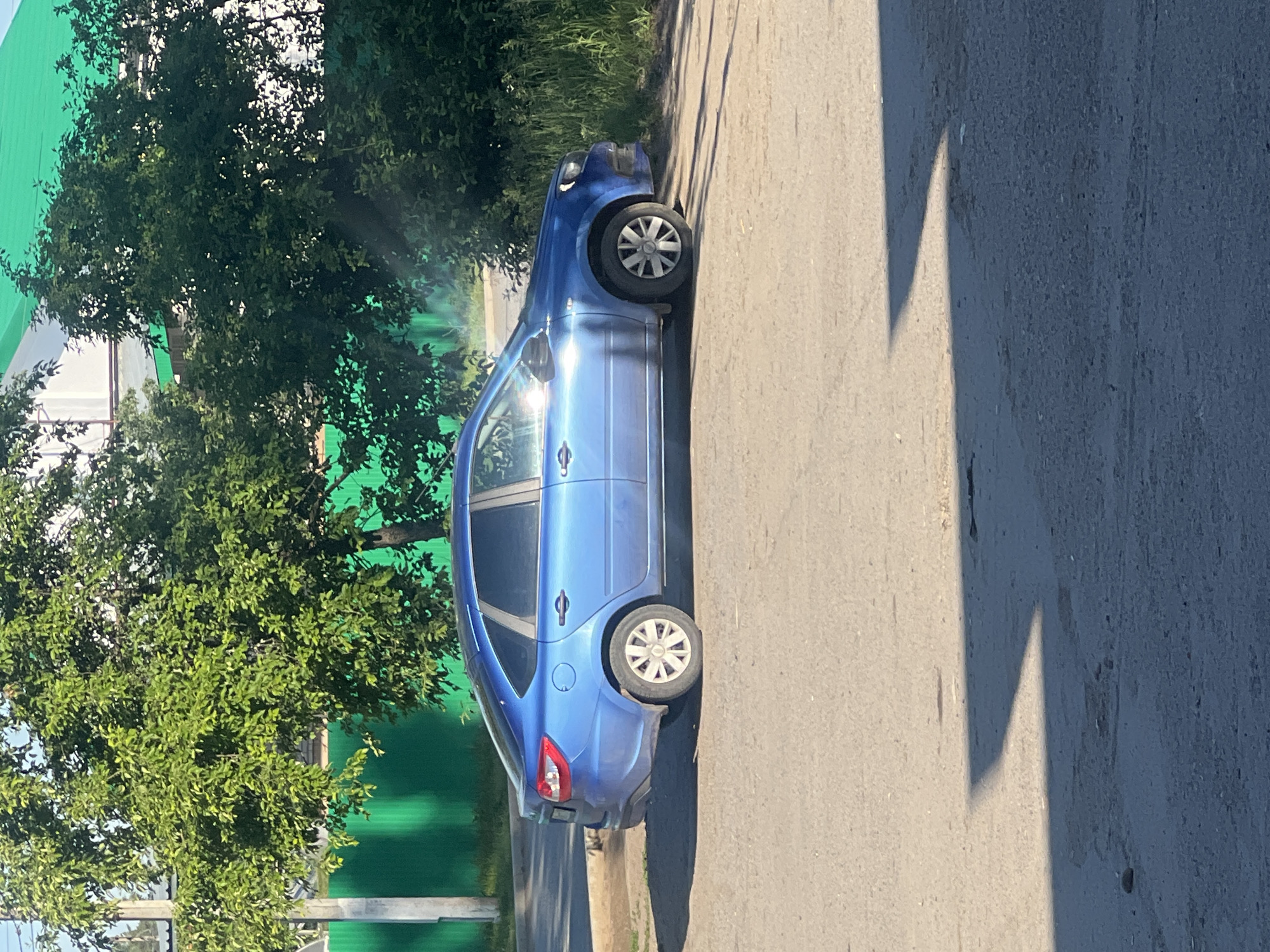 A blue sedan is parked on a paved road in Noviy, Russia. The car is a light blue color and appears to be a sedan. It is parked on the right side of the road, facing left. The road is paved with asphalt and there are some shadows on the ground. In the background, there are trees and a green fence. The sun is shining and the sky is blue.