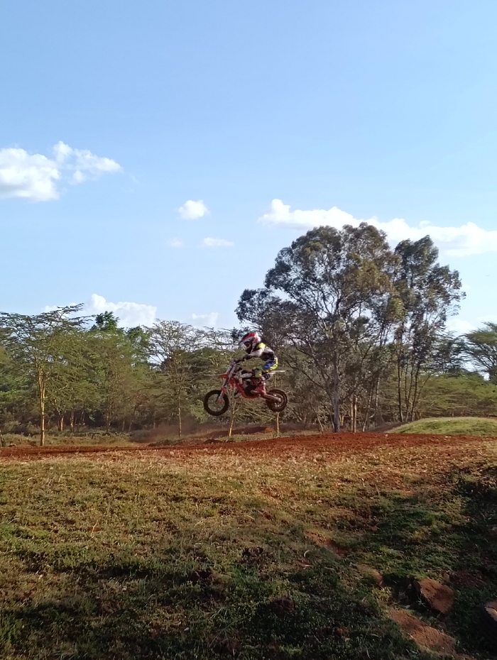 A motocross rider is depicted mid-air on a small-scale red dirt bike, having launched from an earthen ramp on a reddish-brown dirt track. The rider, positioned centrally, wears a multi-colored full-face helmet, goggles, a white, black, yellow, and red long-sleeved jersey, and dark pants. The dirt bike's wheels are off the ground, with visible suspension compression. The foreground comprises a grassy slope interspersed with patches of reddish-brown soil and dark rocks. The midground features the motocross track composed of reddish-brown earth, showing a significant jump. The background consists of a dense, varied woodland with numerous trees, some exhibiting sparse, light green foliage and others denser, darker canopies. The sky is clear blue with scattered white cumulus clouds. This scene is consistent with a dedicated off-road or motocross track situated within a natural, bushland environment on the outskirts of Nairobi, Kenya, such as the Jamhuri Race Track area.