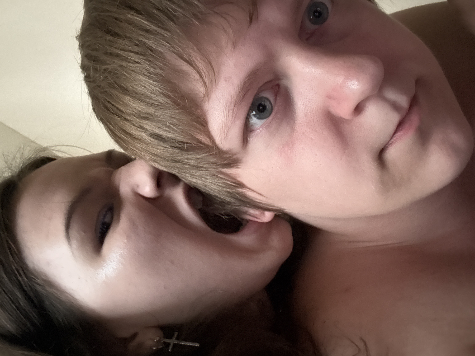 The image is a close-up, intimate portrait of two individuals. In the foreground, a person with light brown hair and blue eyes looks directly at the camera. Their face is slightly turned, with the right eye more prominent. To the left and slightly behind, another person with dark brown hair is visible. Their mouth is open wide, and they appear to be biting or nibbling on the ear of the person in front. A silver cross earring is visible on the left ear of this second individual. The background is a plain, light-colored wall, suggesting an indoor setting. No specific location in Saint Petersburg, Russia, can be identified from the image itself.
